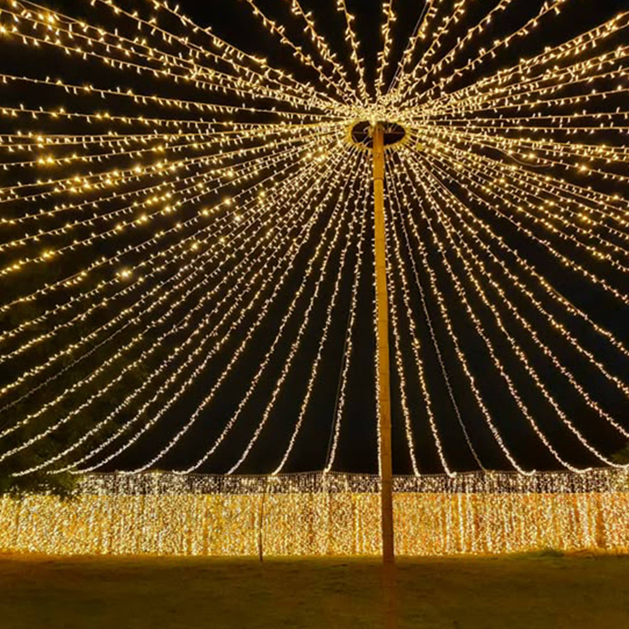 Escena nocturna con luces que irradian de un poste central, asemejando un dosel. Una pared de luces en la parte inferior.