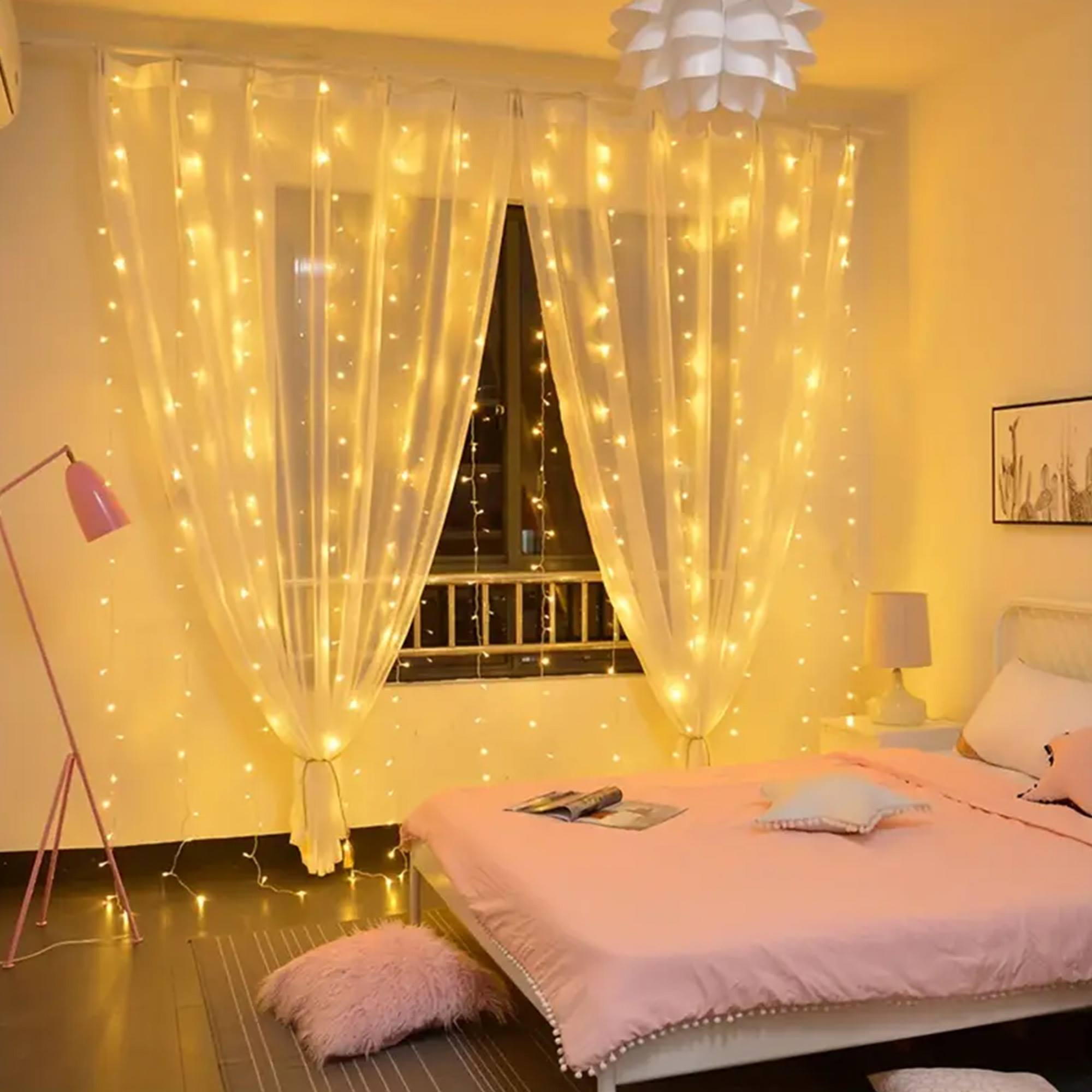 Dormitorio con luces de hadas en cortinas. Cama rosa, lámpara y almohadas. Iluminación cálida.