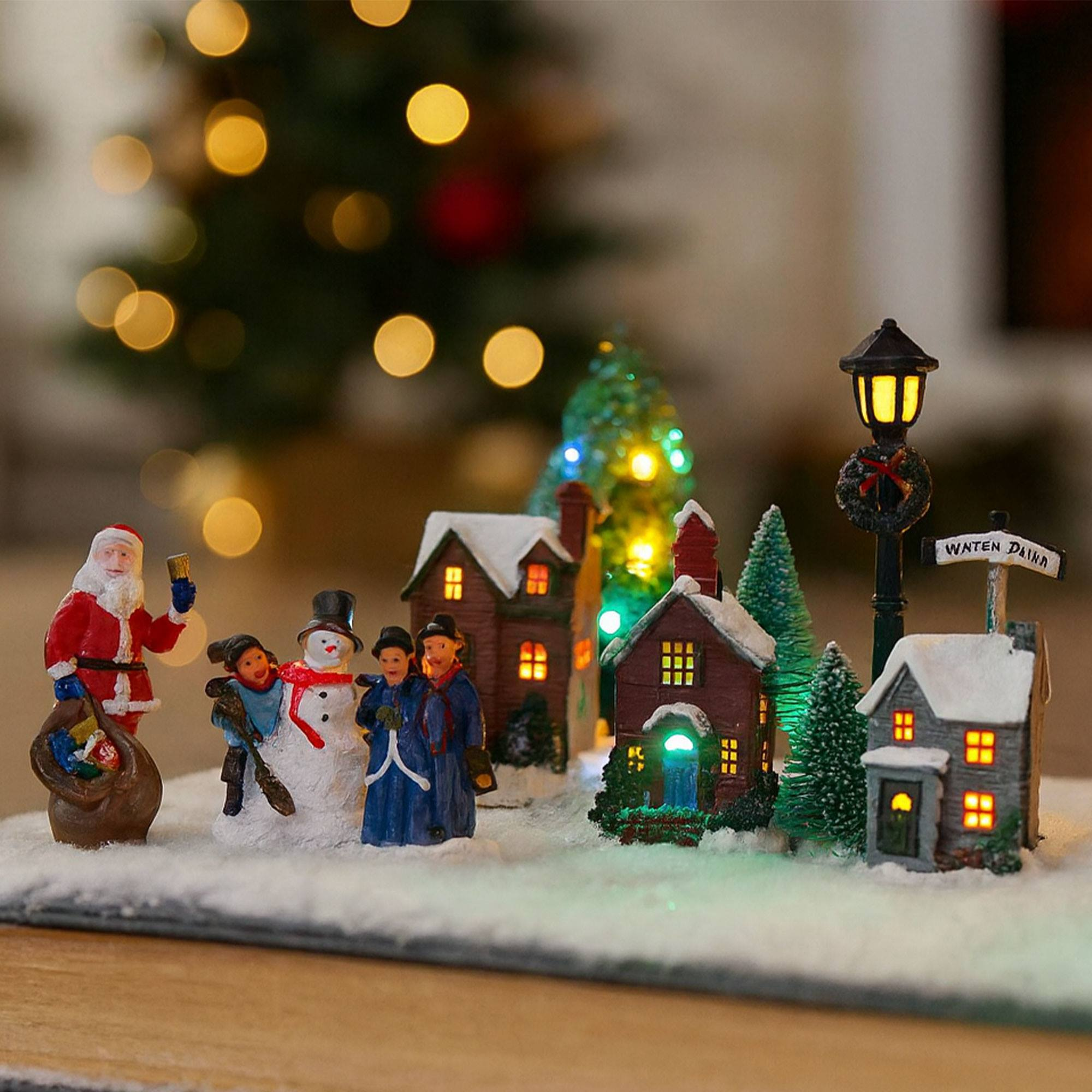 Una escena de pueblo navideño con Papá Noel, muñeco de nieve, figuras, casas, árboles y farola, todo sobre nieve.
