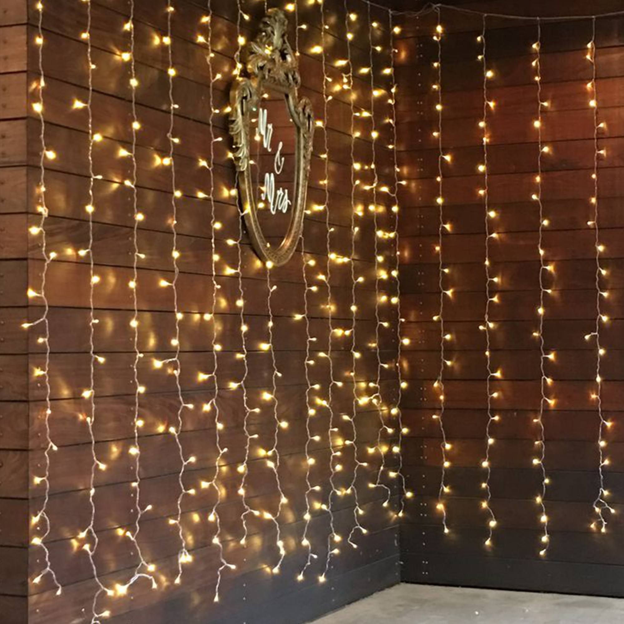 Una pared de madera decorada con guirnaldas de luces cálidas. Un espejo vintage con 'Mr & Mrs' cuelga.
