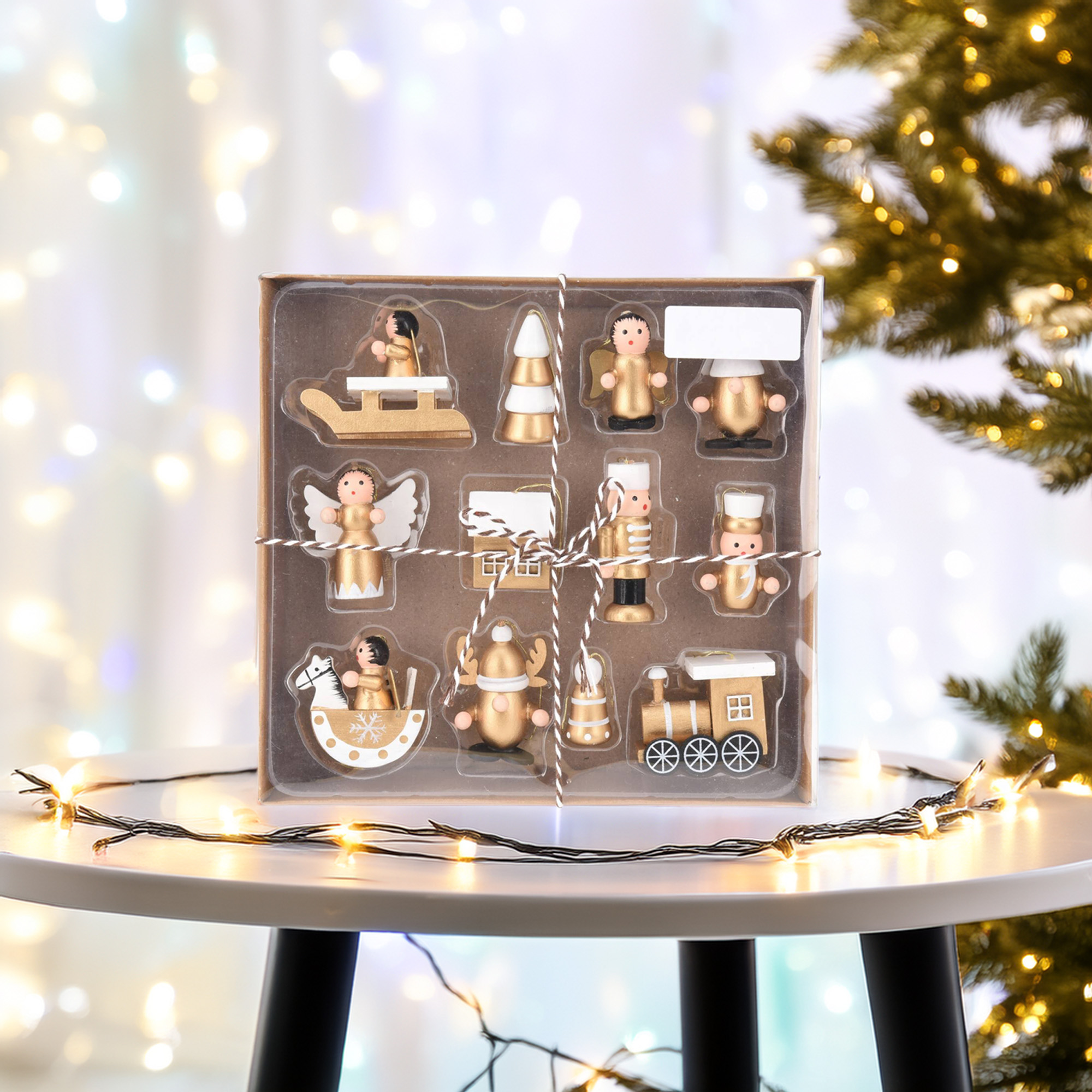 Un conjunto de adornos navideños de madera, que incluyen ángeles y un tren, sobre una mesa blanca, con luces de hadas.