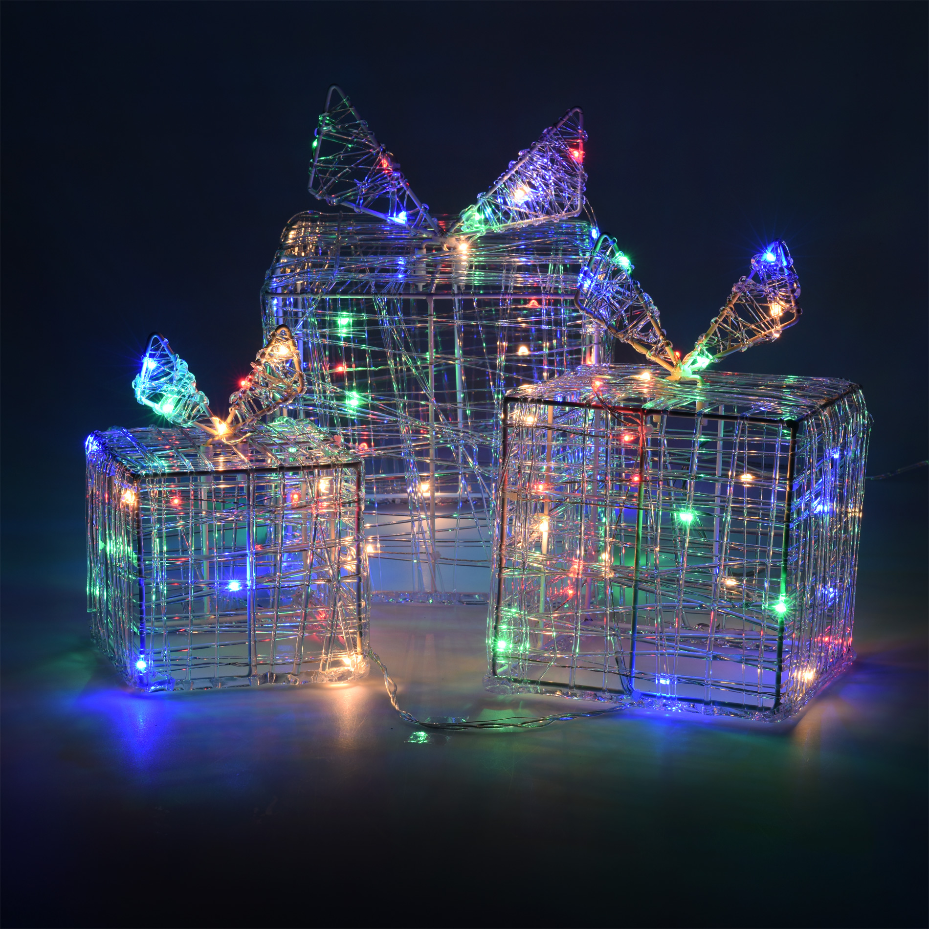 Tres cajas de regalo con estructura de alambre y luces de colores. La más grande tiene un lazo. Fondo oscuro.