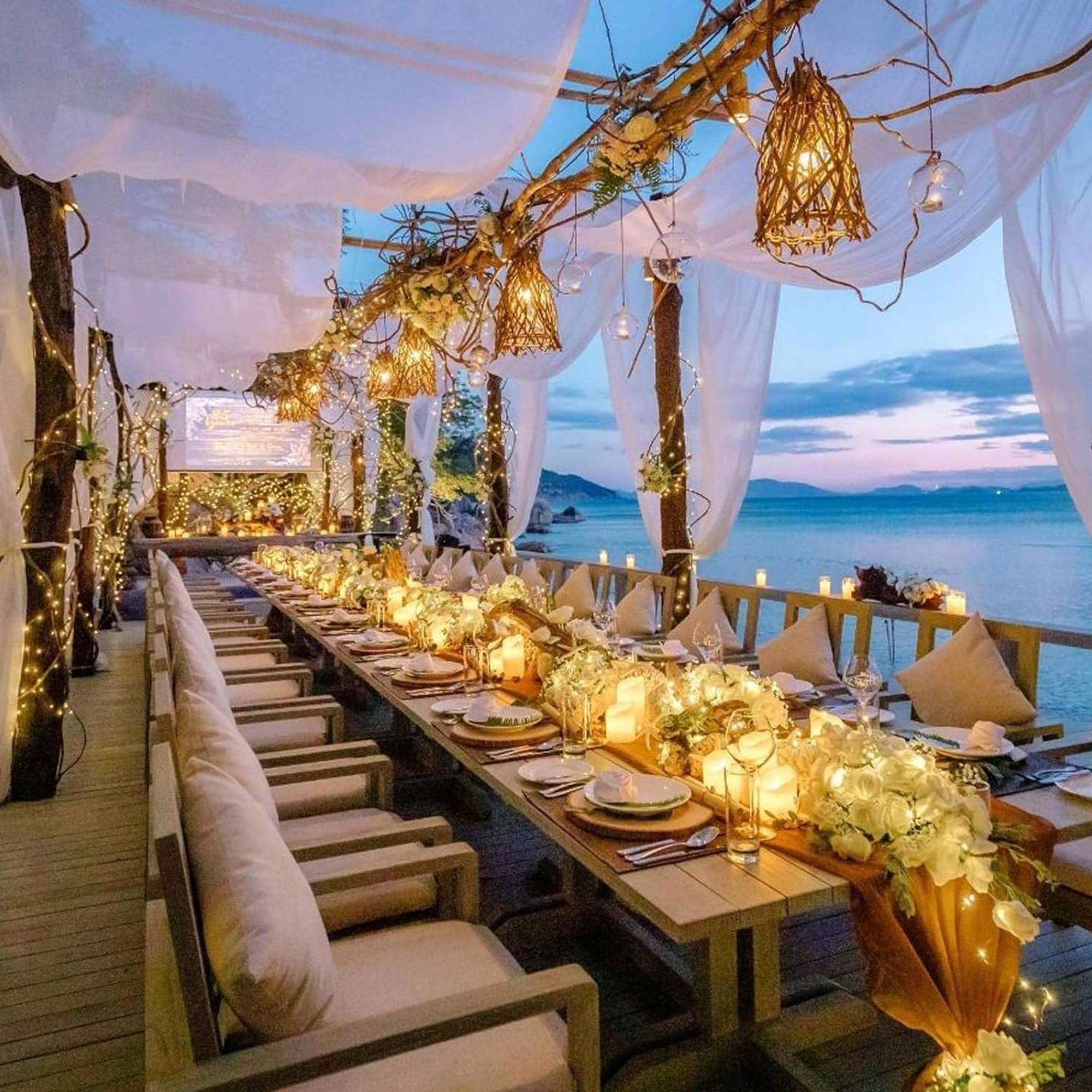 Una mesa de cena bellamente decorada en una terraza con vista al mar al anochecer, decorada con luces y flores.