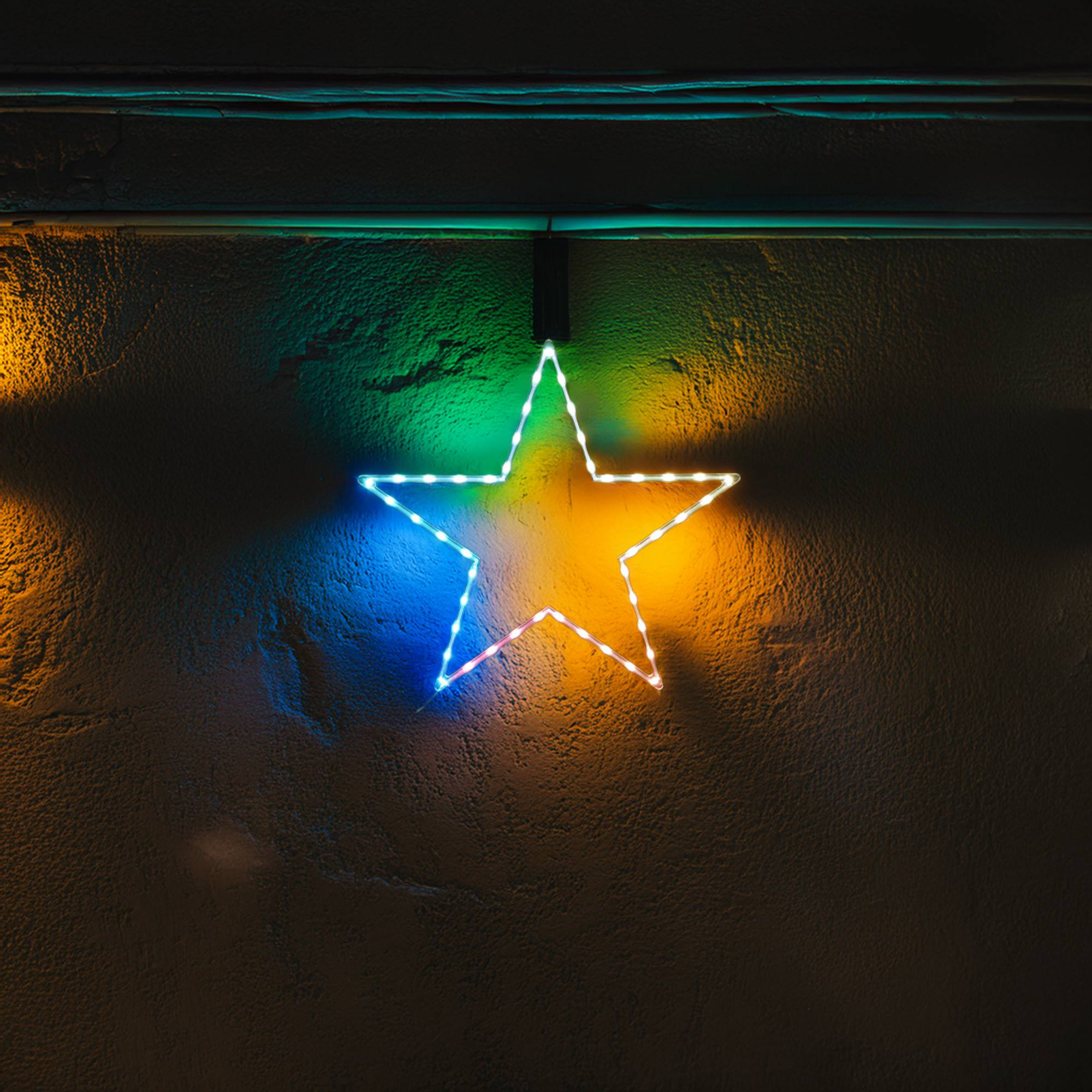 Una luminaria en forma de estrella brillante con colores neón cuelga de una pared oscura texturizada. La estrella tiene luces azules, verdes y amarillas.
