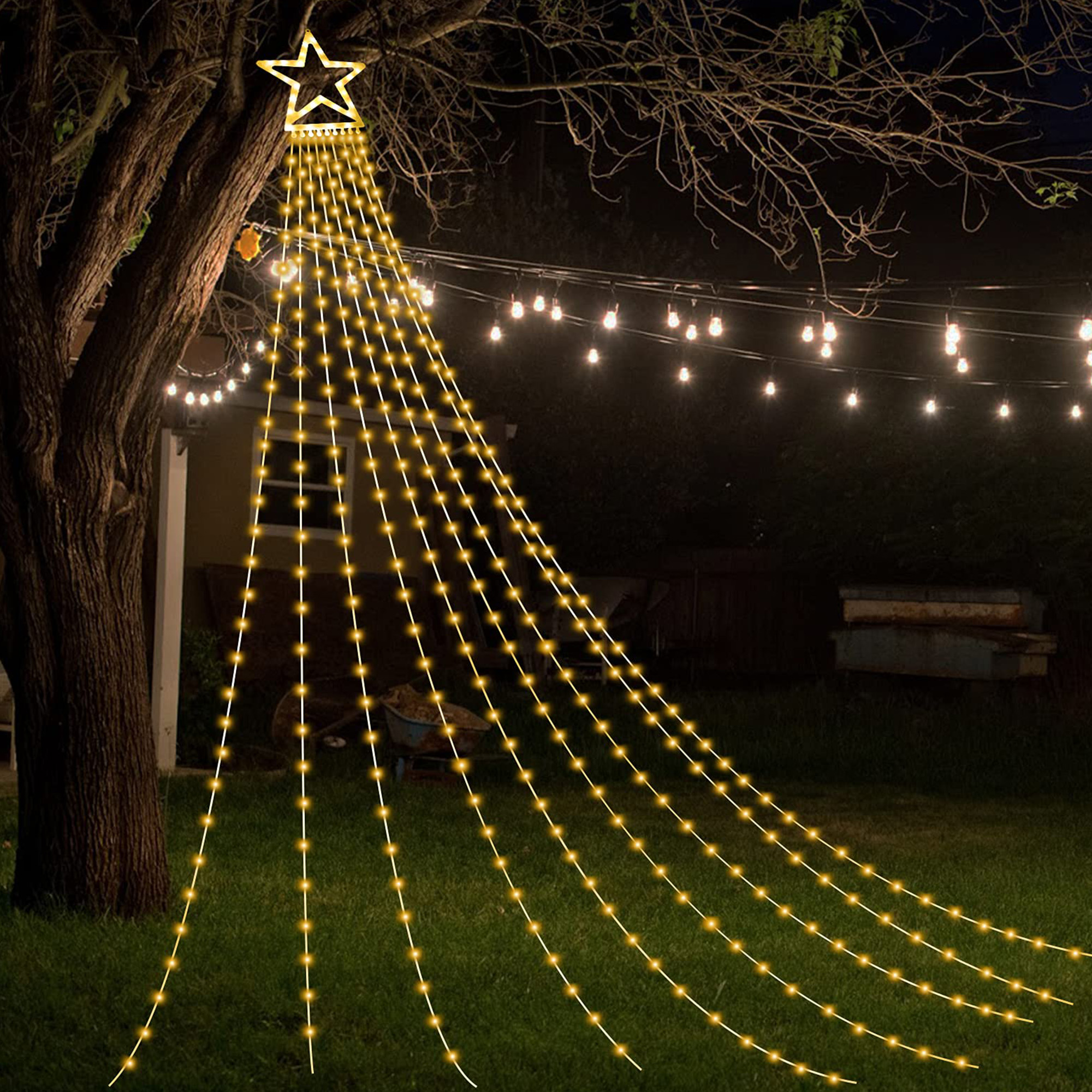 Una lámpara en forma de estrella cuelga de un árbol, con hilos de luces que caen sobre la hierba. Luces de cadena en el fondo.
