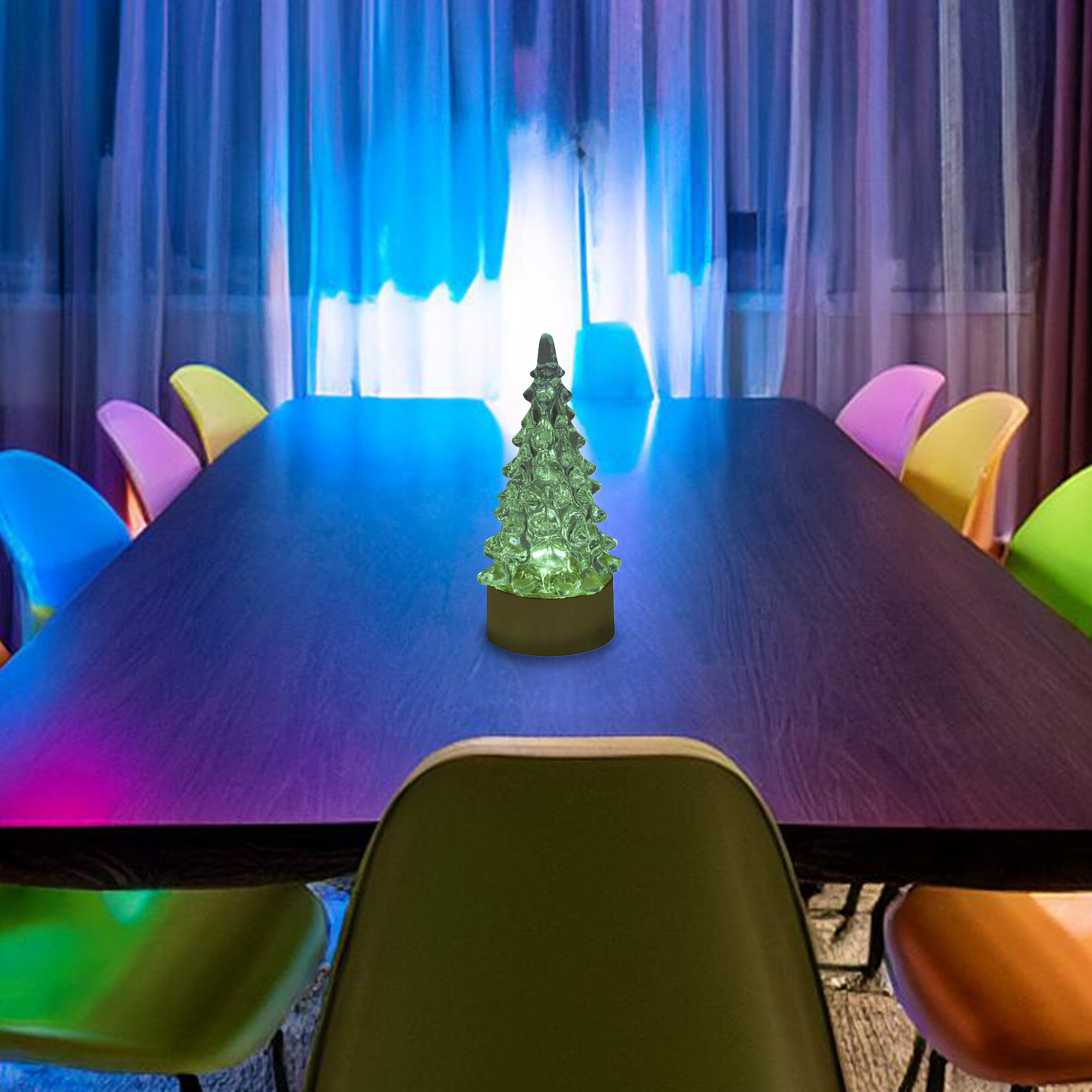 Un árbol de Navidad decorativo e iluminado está sobre una mesa rodeado de sillas coloridas, una cortina azul claro al fondo.