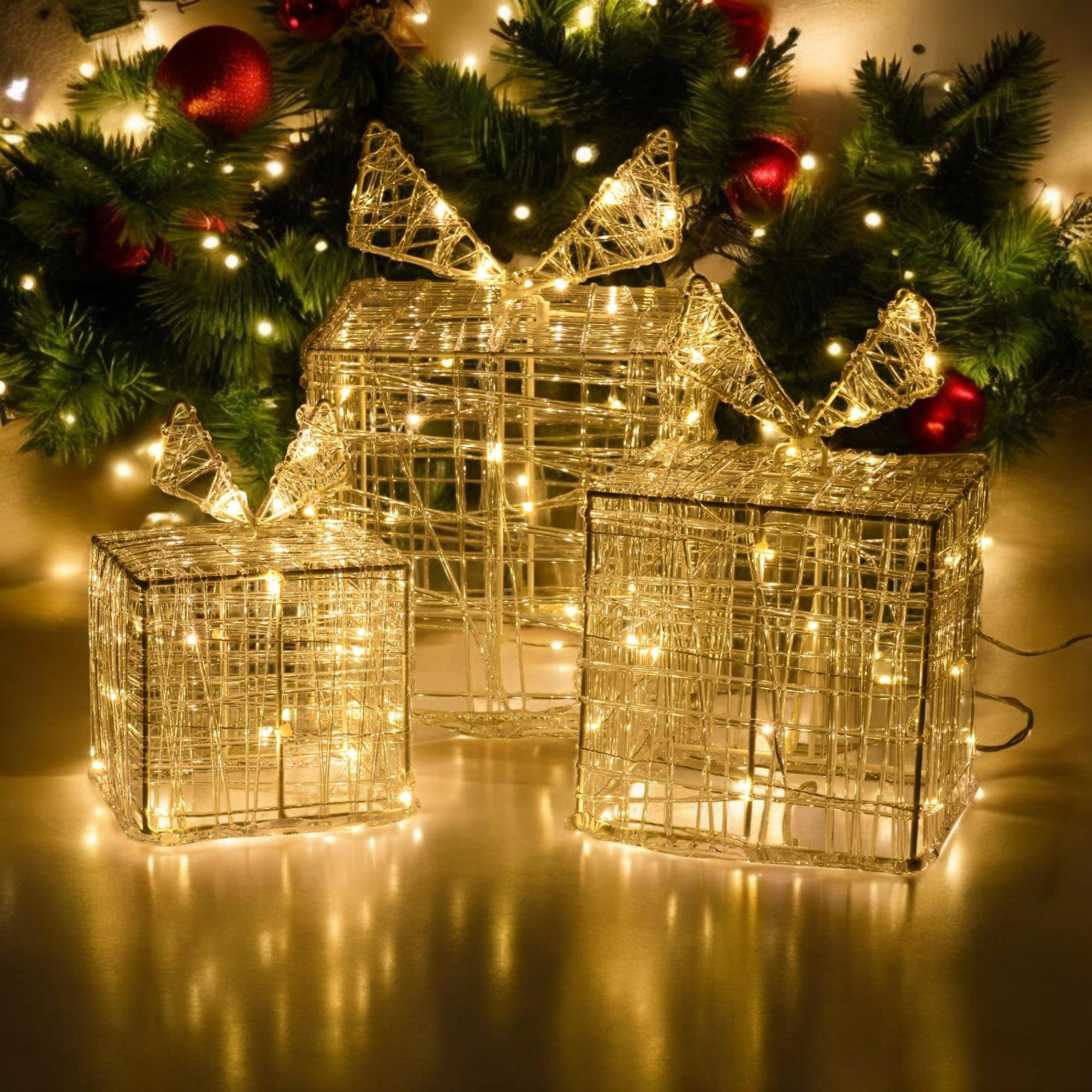 Tres cajas de regalo de alambre iluminadas con lazos frente a un árbol de Navidad con adornos rojos.