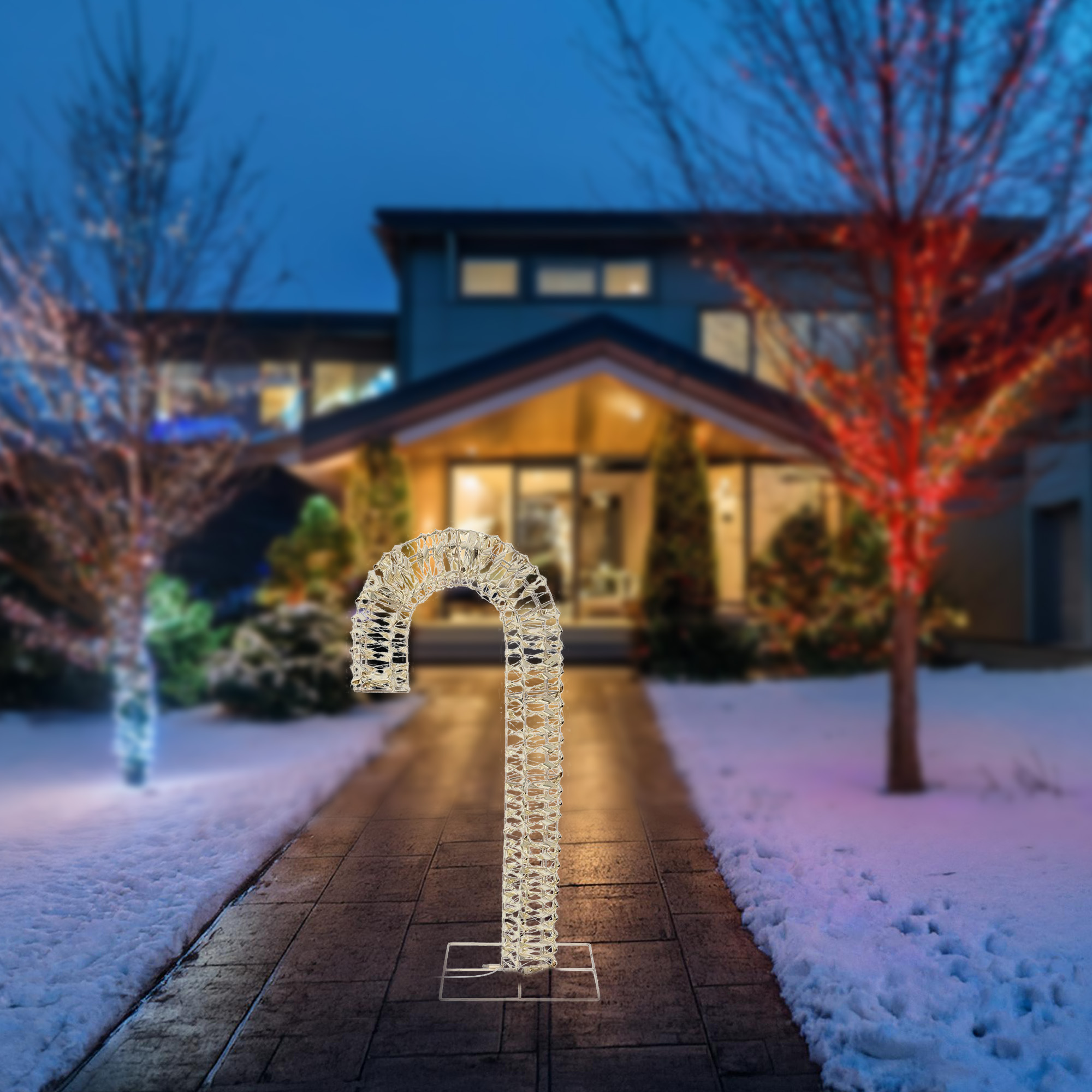 Una decoración de bastón de caramelo iluminada en un camino nevado conduce a una casa al anochecer. Los árboles están decorados con luces.