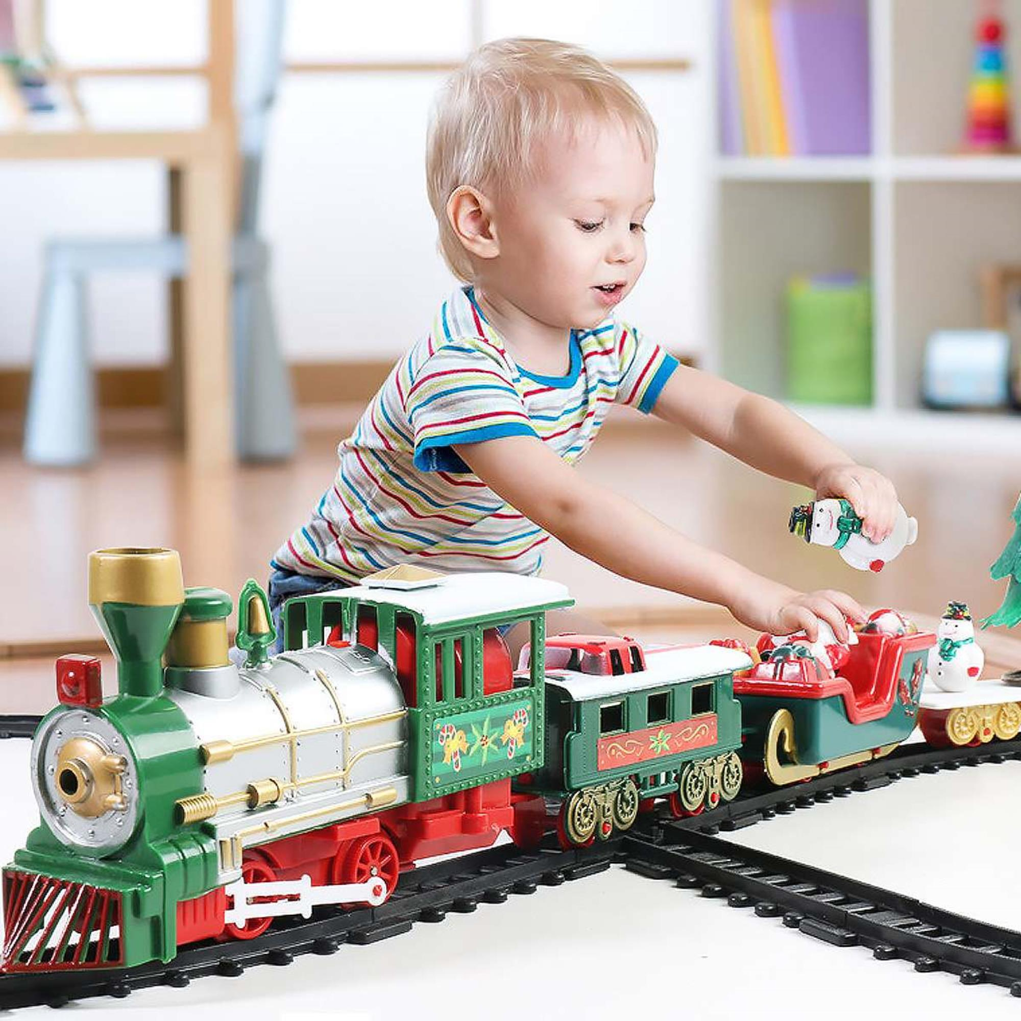 Un niño pequeño juega con un tren de juguete. El tren es verde, rojo y plateado, sobre una vía negra. El niño sostiene un juguete.