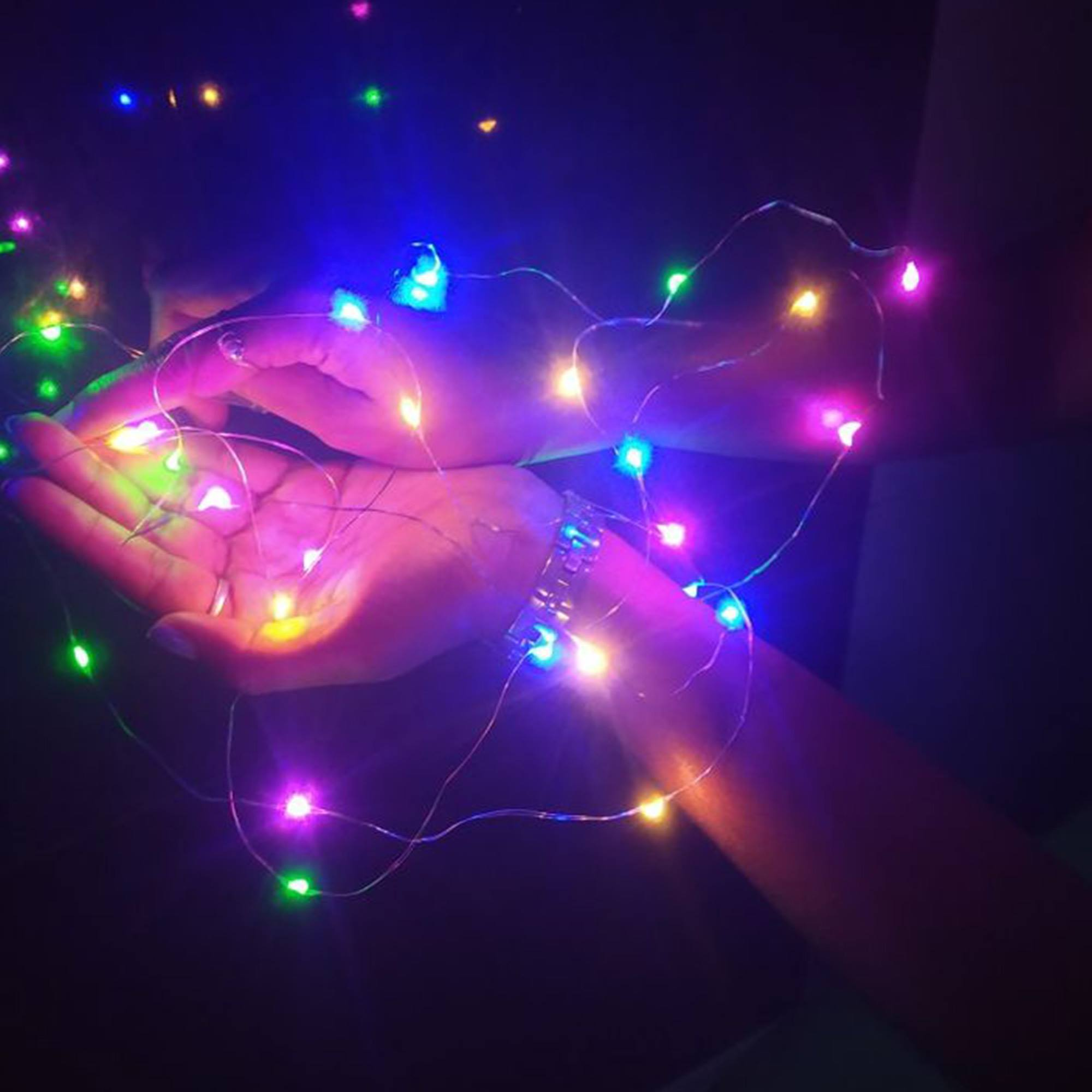 Manos sosteniendo luces de cadena de varios colores. Una pulsera plateada está en la muñeca. Fondo oscuro.