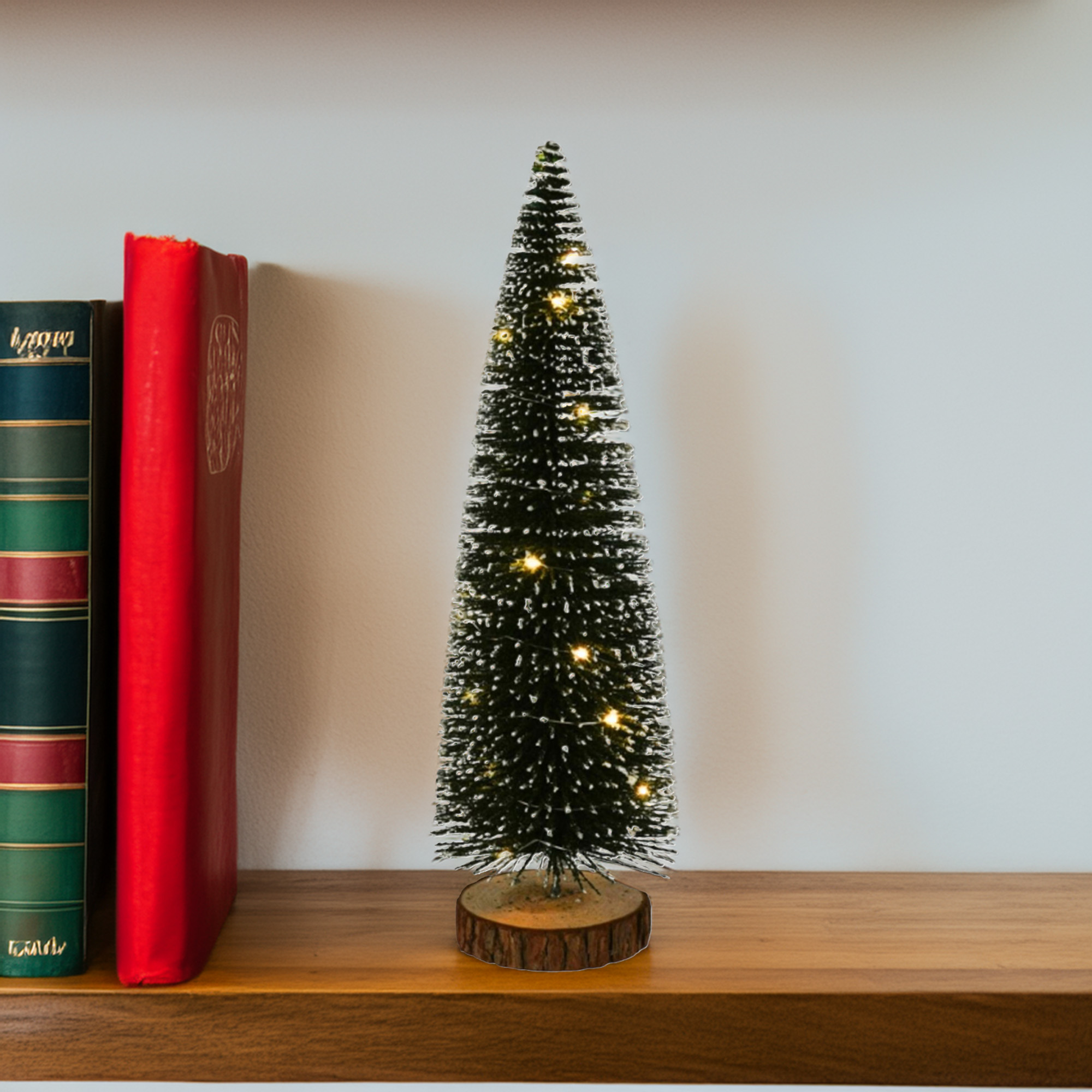 Un pequeño árbol de Navidad con luces está en una estantería de madera. Dos libros a la izquierda, uno rojo y otro verde.