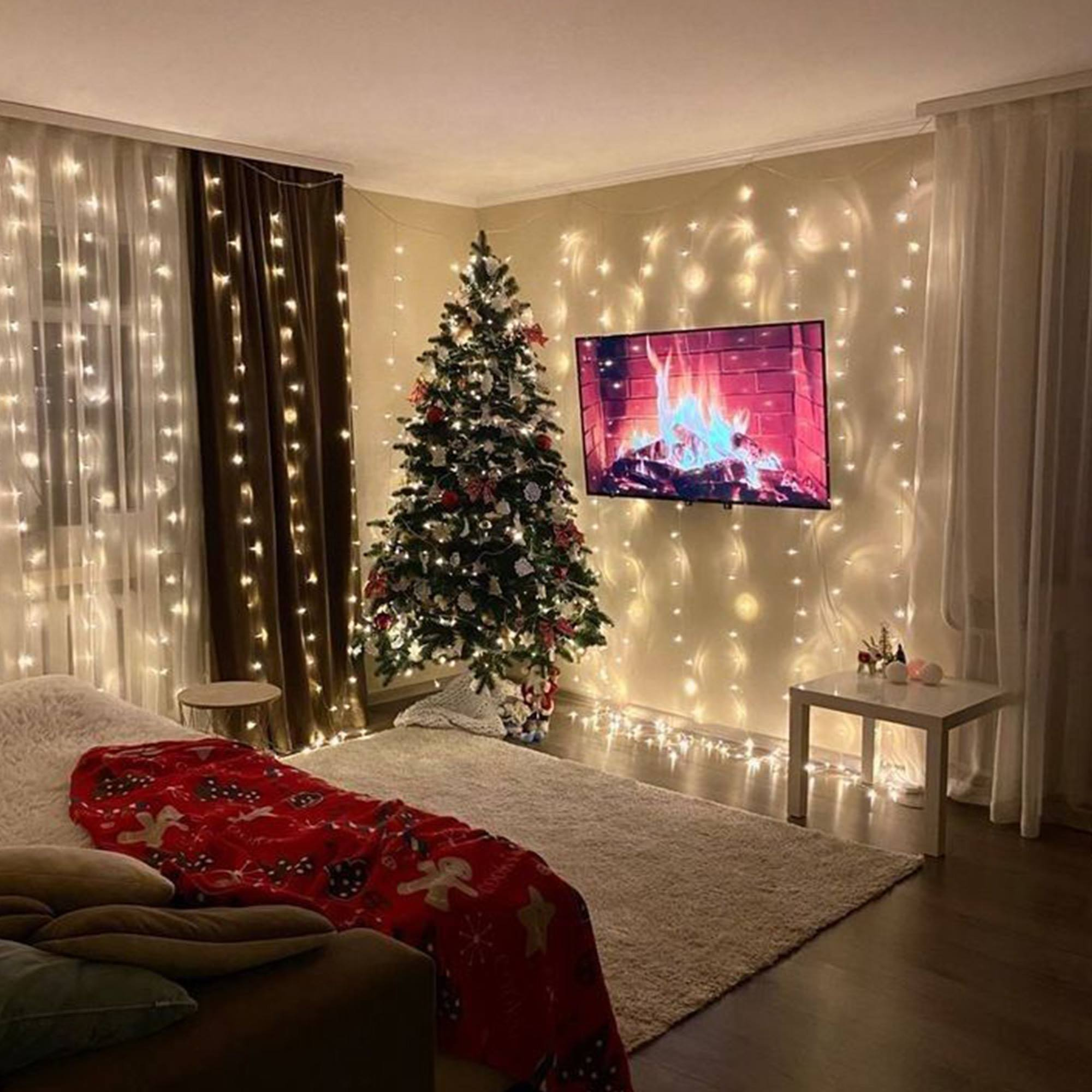 Un árbol de Navidad decorado en una habitación con luces de hadas. Un televisor muestra una chimenea. Manta roja en el sofá.