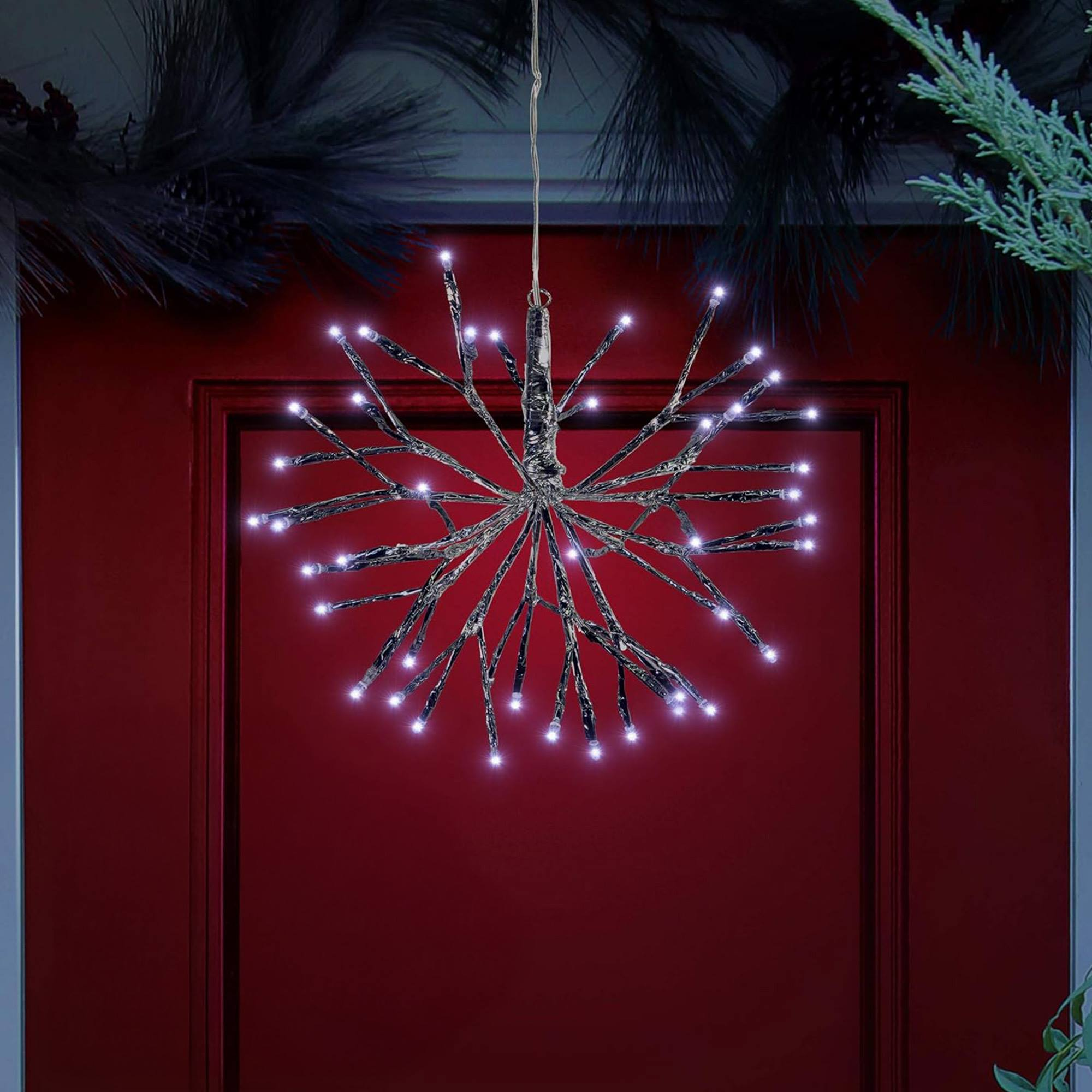 Una decoración en forma de estrella con luces blancas cuelga frente a una puerta roja. Hay vegetación oscura y piñas arriba y abajo.
