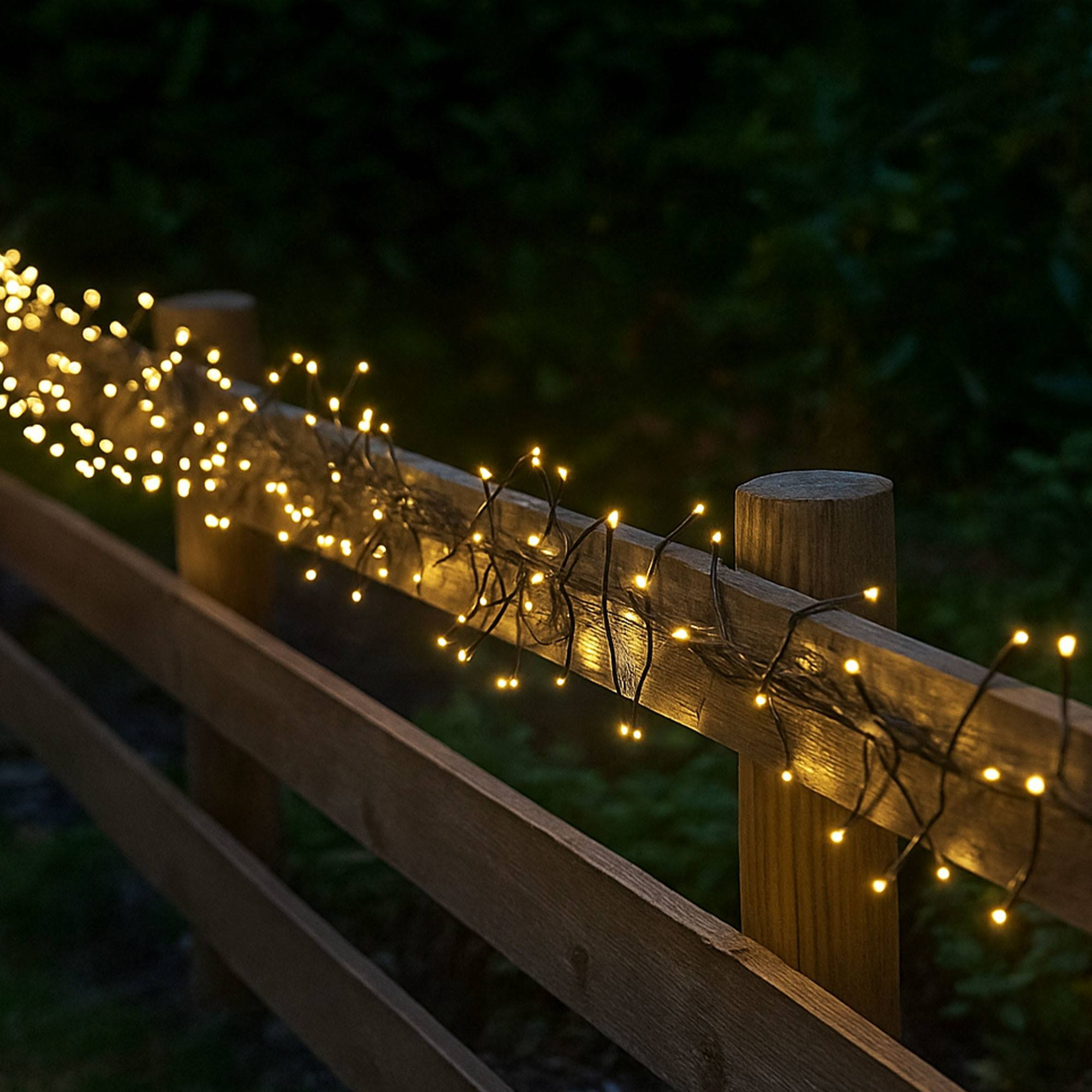 Valla de madera adornada con luces de cadena brillantes. Follaje verde oscuro en el fondo. Escena iluminada por la noche.