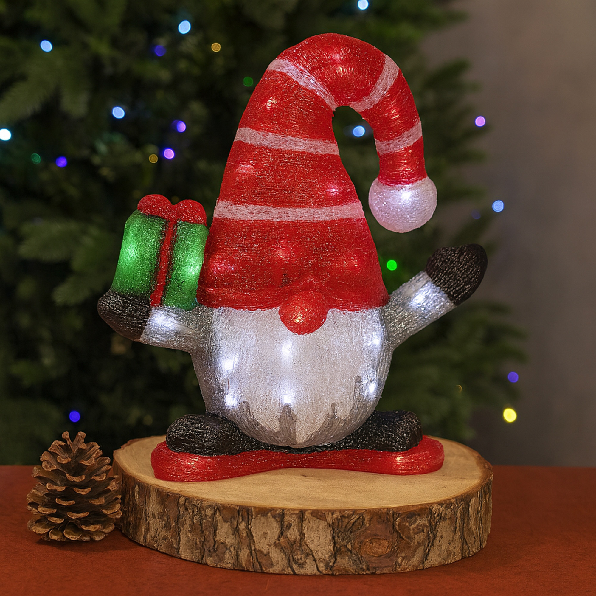 Un gnomo navideño con gorro rojo y regalo, sobre una base de madera. Una piña y un árbol de Navidad en el fondo.