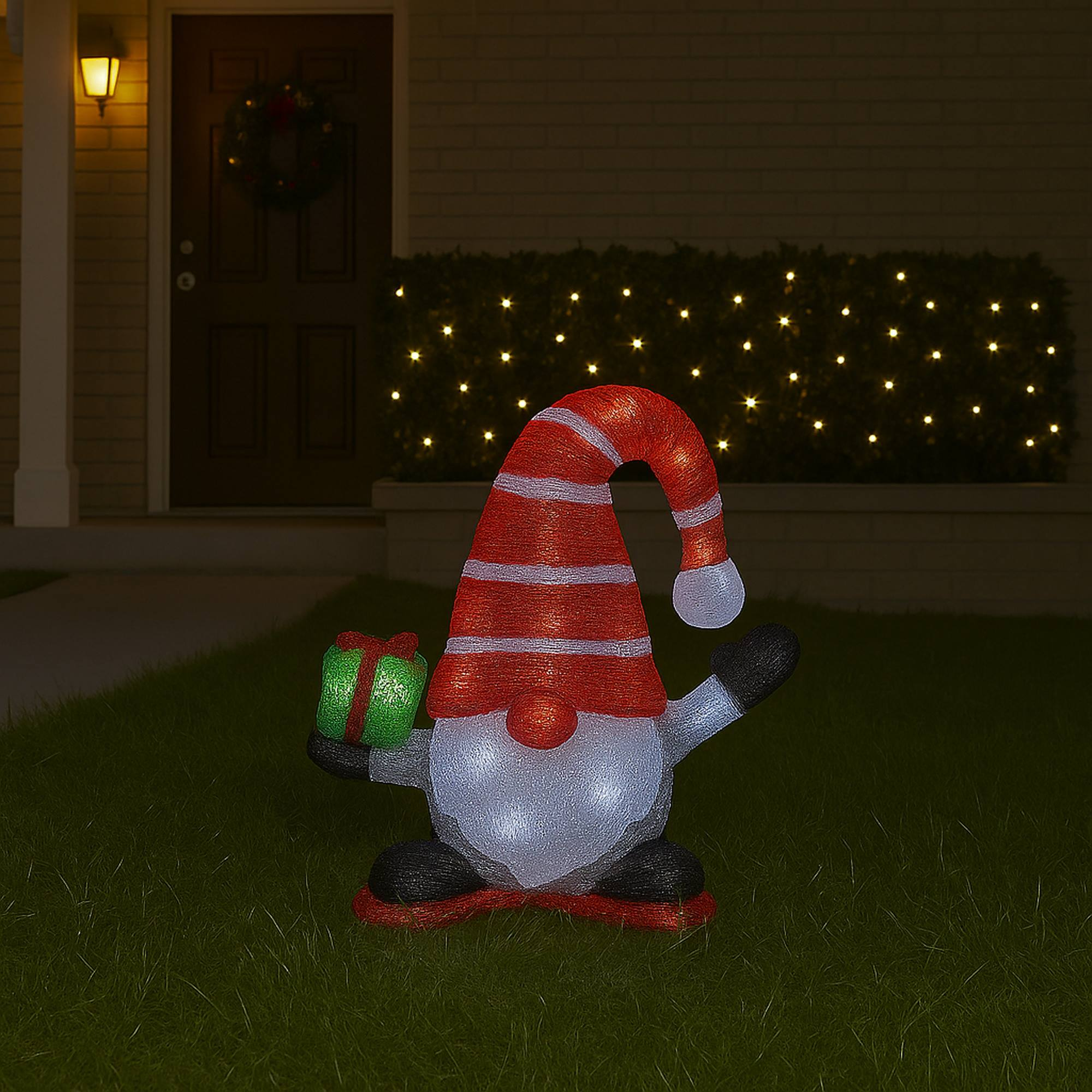 Un gnomo de jardín con un gorro rojo y un regalo verde está sobre la hierba. Una casa y luces navideñas al fondo.