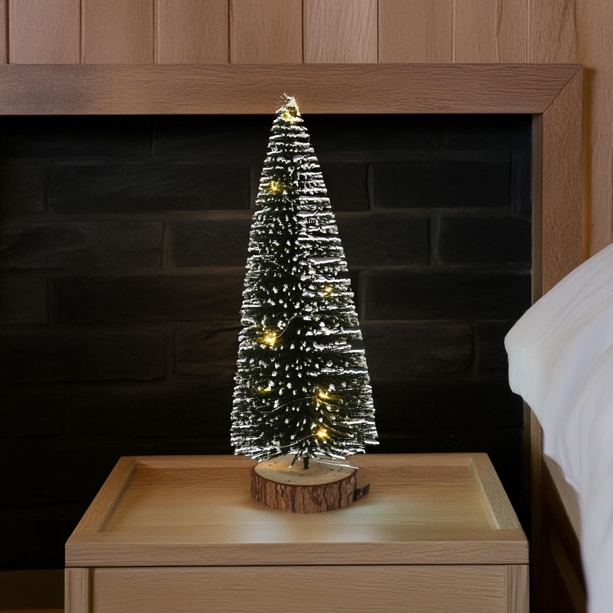 Un pequeño árbol de Navidad con luces está en una mesita de noche. Una cama está a la derecha. Pared de ladrillo al fondo.