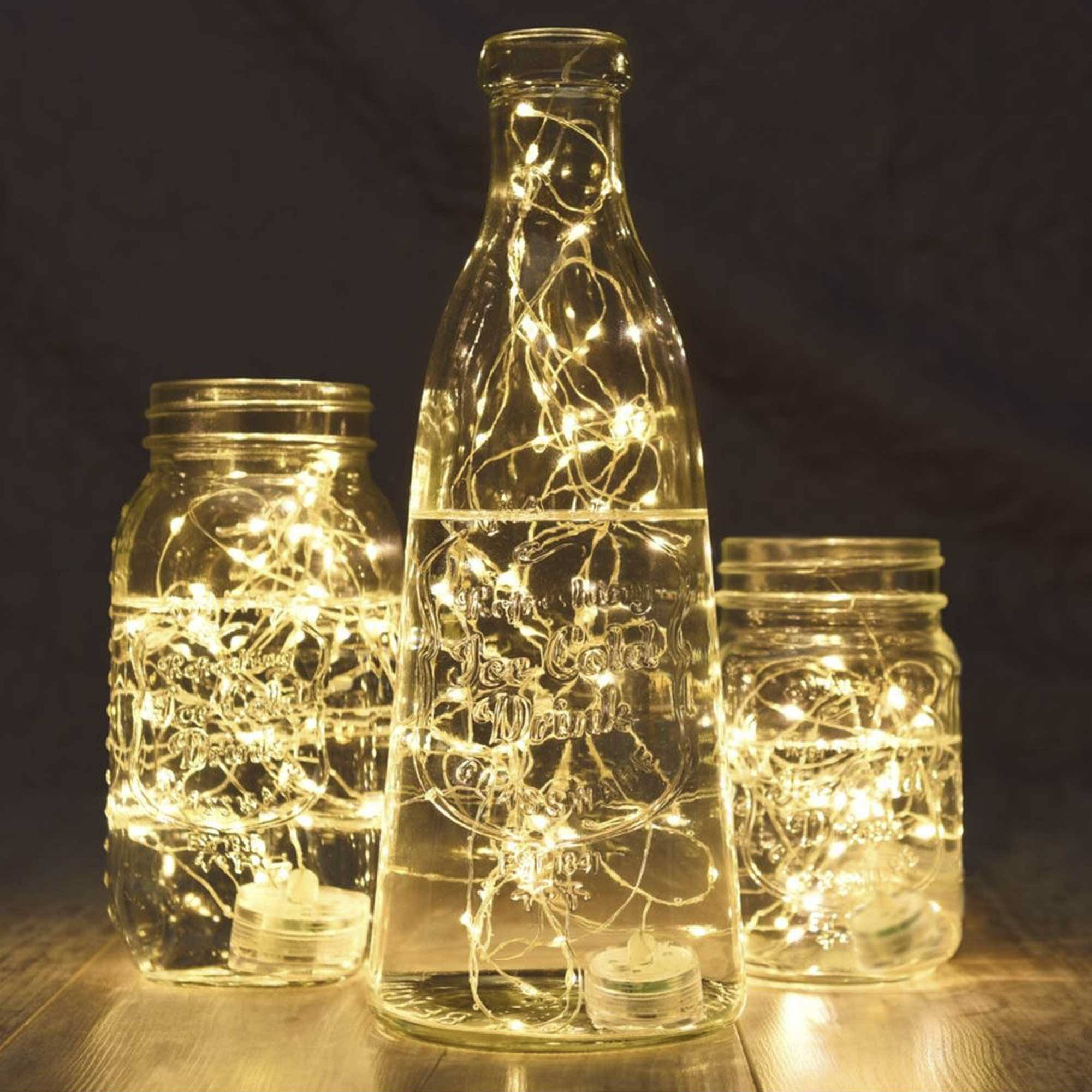 Tres frascos de vidrio con luces de hadas. El frasco central tiene forma de botella. Frascos sobre madera, fondo oscuro.