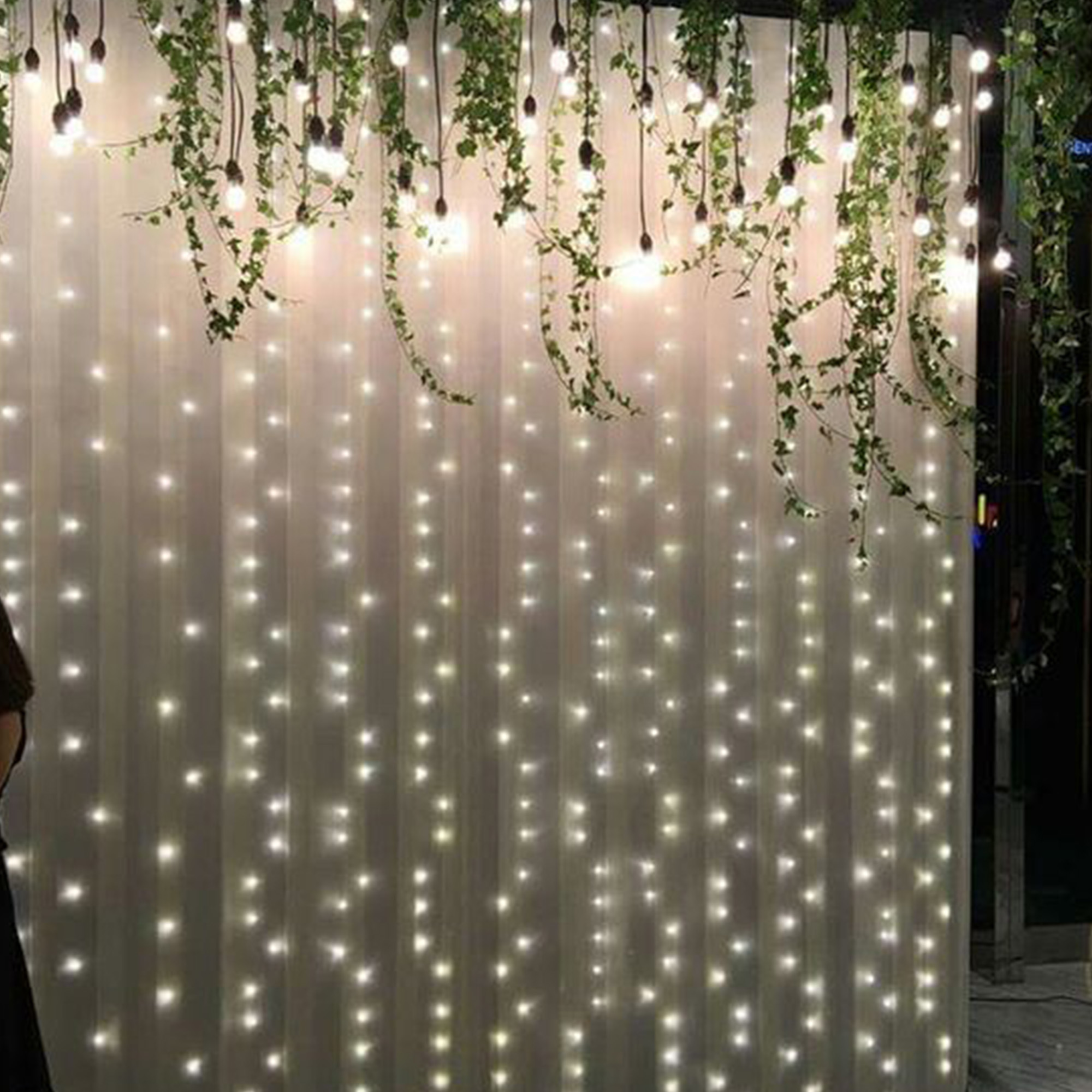 Un telón de fondo blanco con luces de hadas colgantes y vegetación. Las luces son cálidas y la escena es elegante.