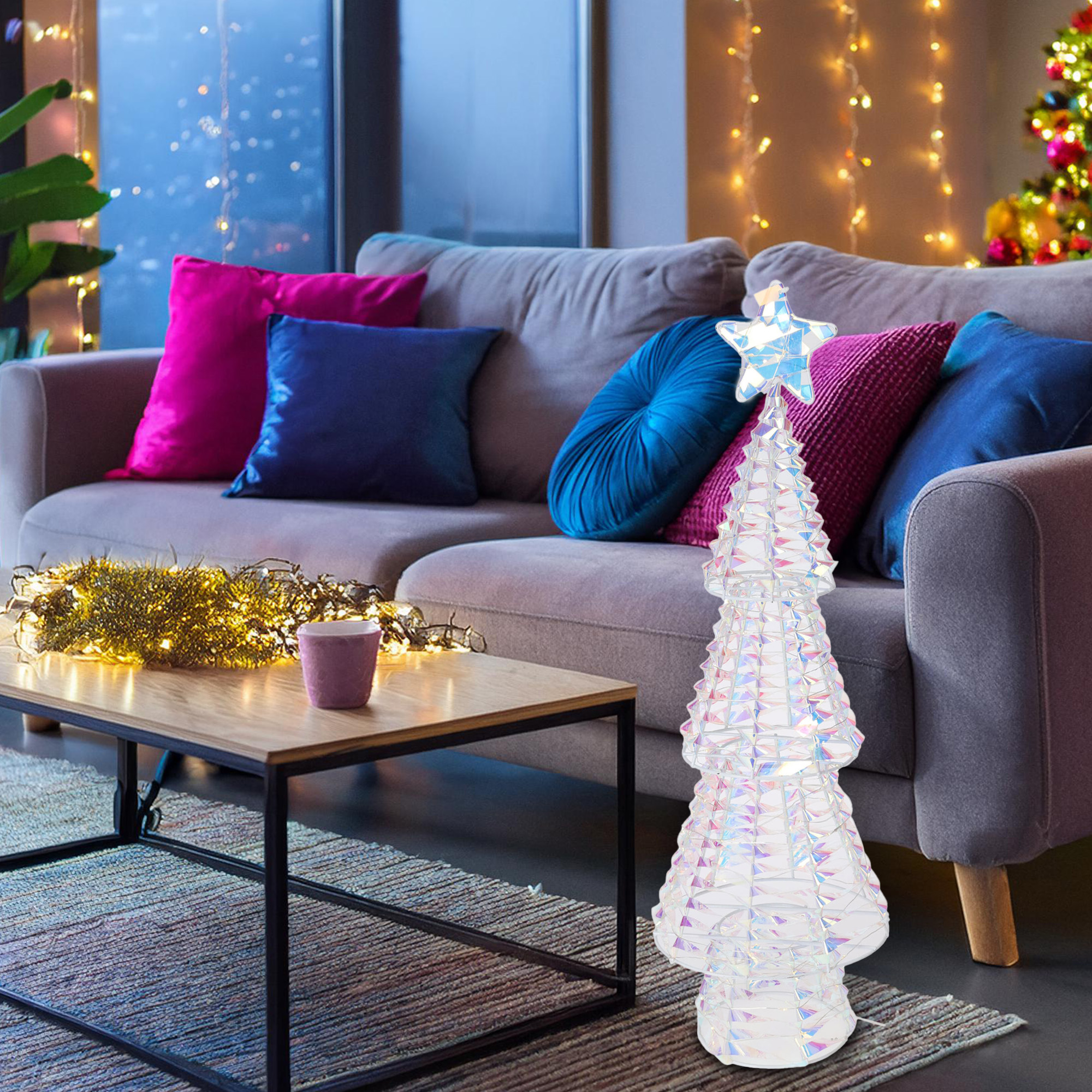 Un árbol de Navidad de cristal iluminado se encuentra frente a un sofá con almohadas. Una mesa de centro con luces y una taza está al frente.