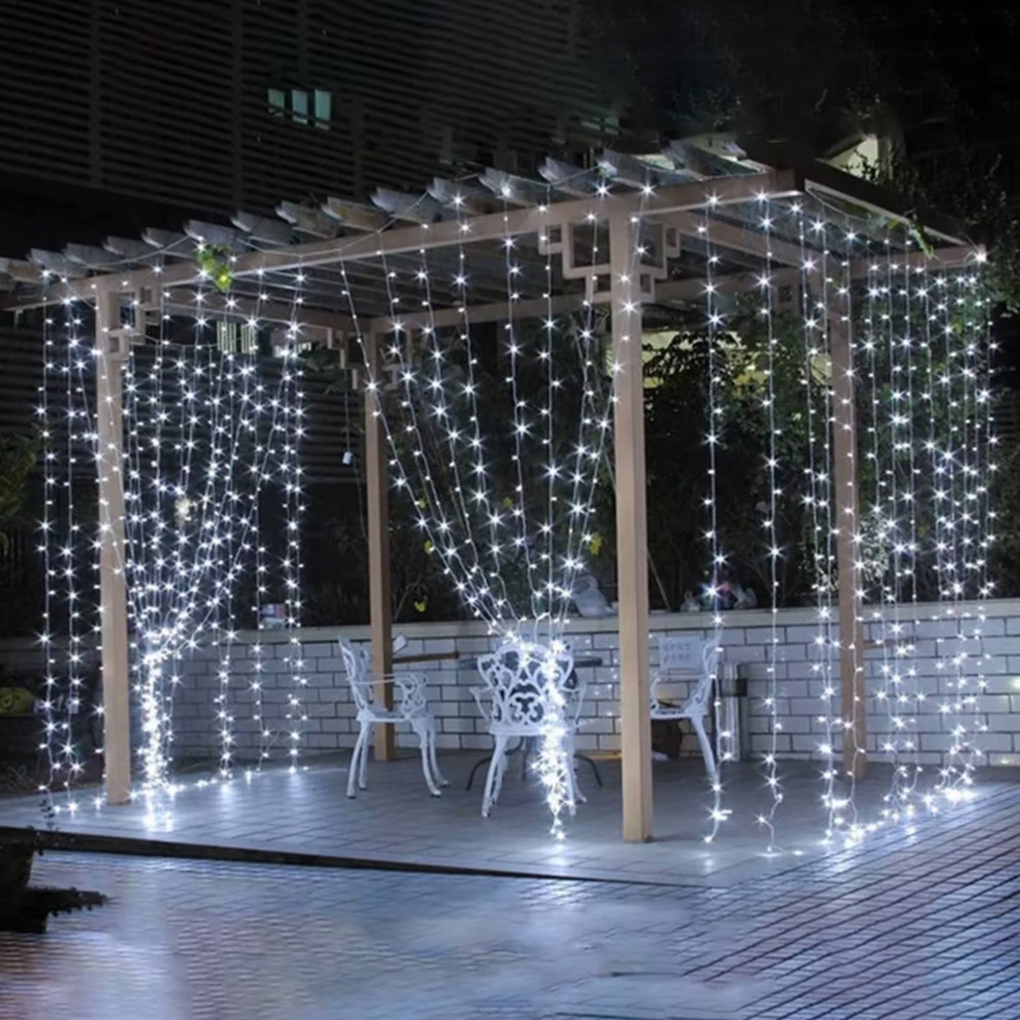 Una pérgola iluminada con luces de cadena blancas. Cuatro sillas blancas están debajo de la pérgola. Fondo oscuro con edificio.