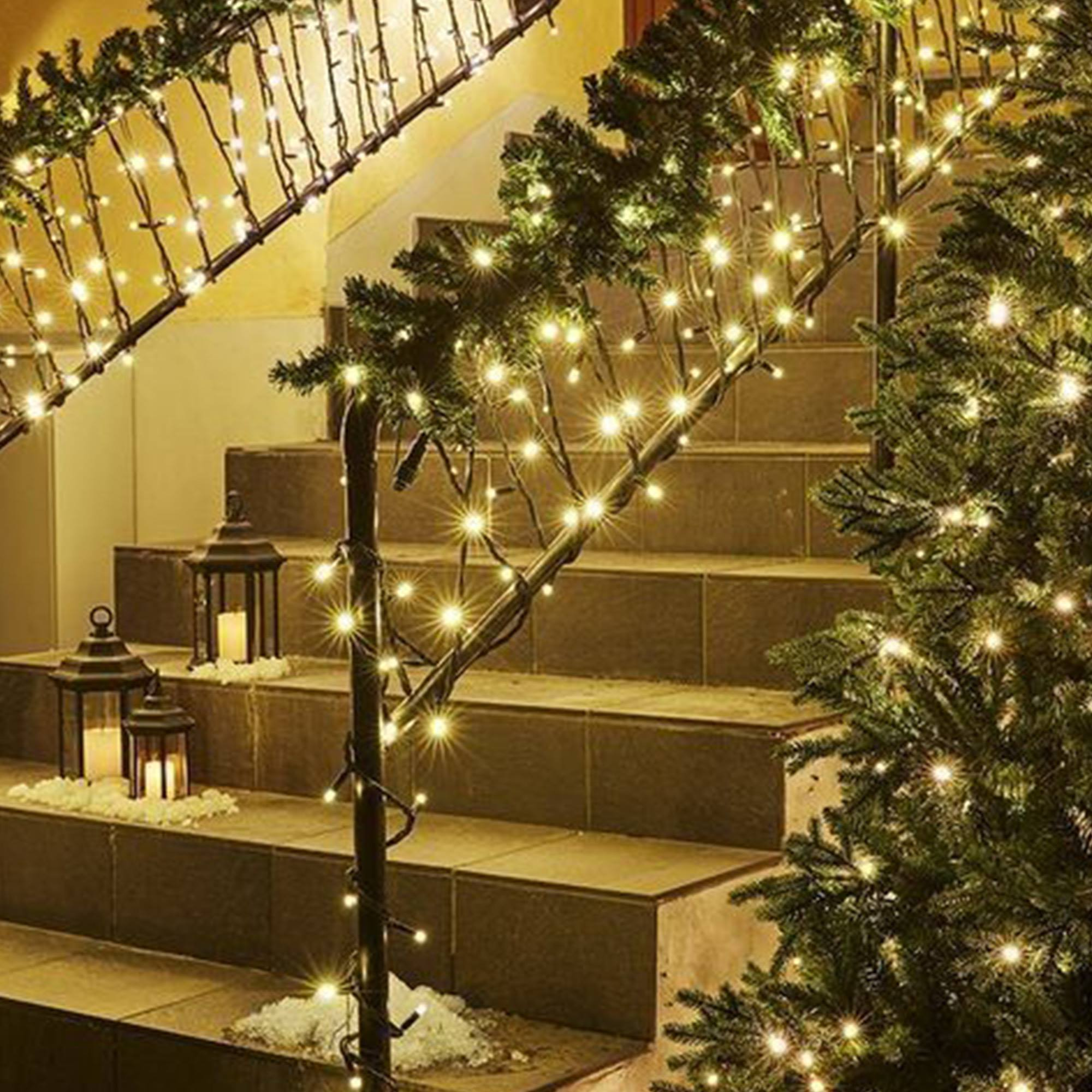 Una escalera decorada para Navidad con luces. Faroles con velas sobre los escalones. Un árbol de Navidad a un lado.