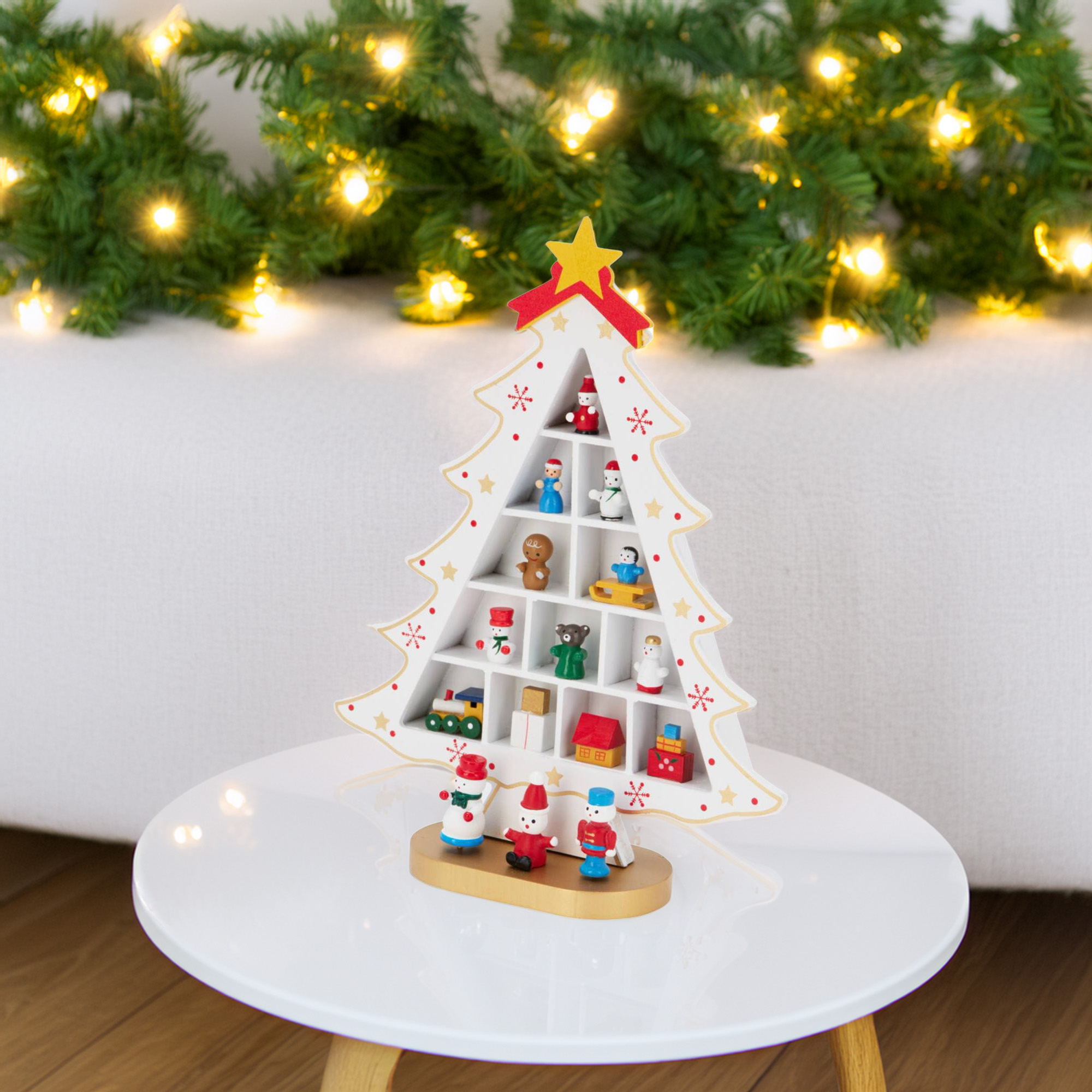 Un estante blanco con forma de árbol de Navidad contiene pequeñas figuras. Está sobre una mesa blanca. Luces de hadas y vegetación al fondo.