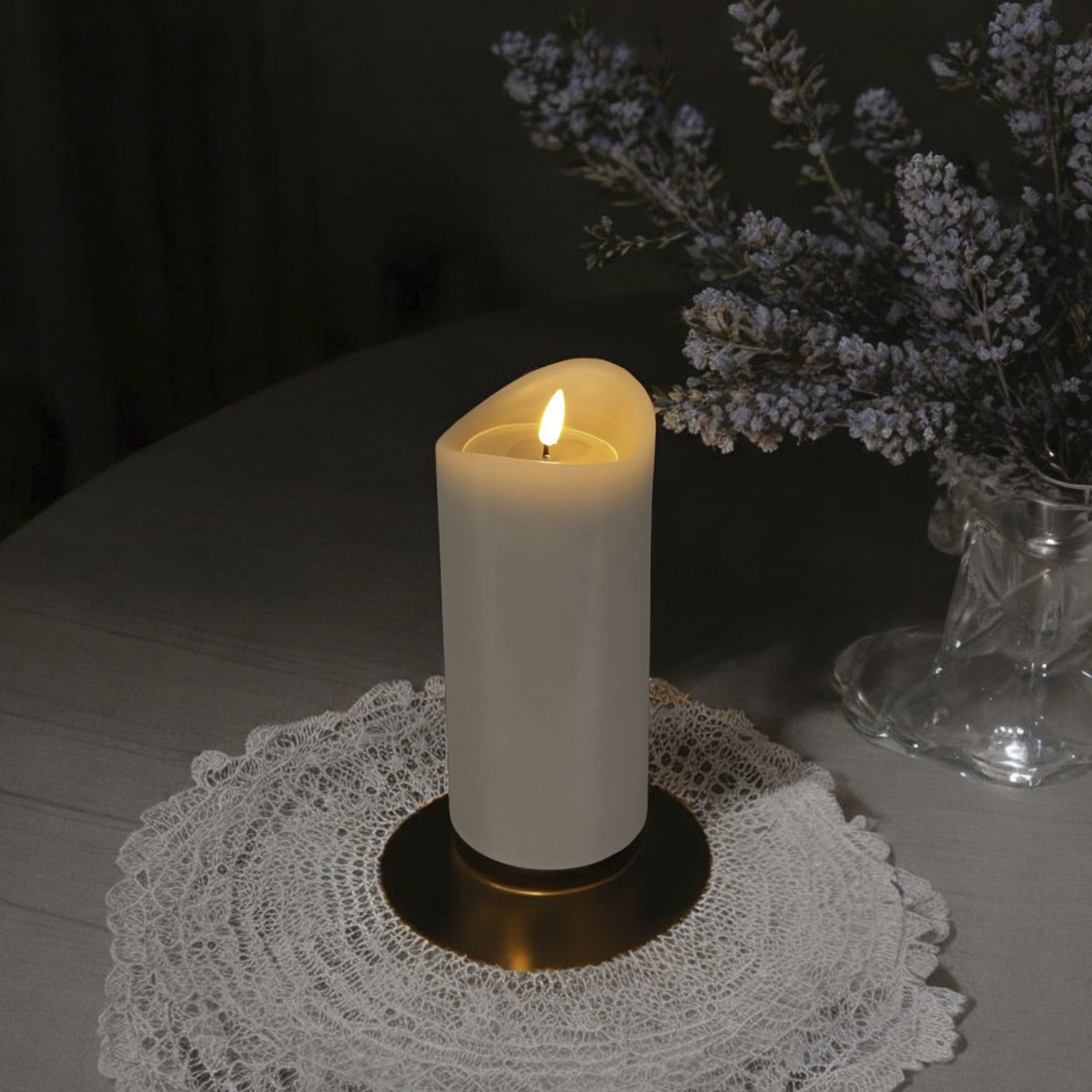 Una vela blanca encendida sobre un tapete de encaje, con flores en un jarrón. Fondo oscuro con una mesa.