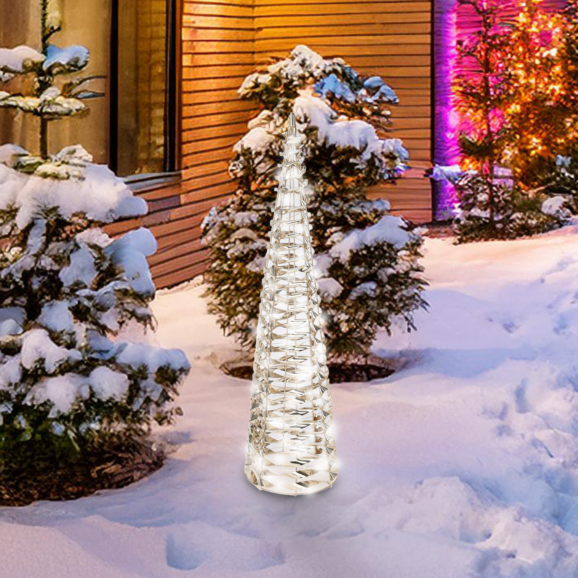 Un árbol decorativo iluminado en forma de cono se alza en la nieve. Árboles, una casa y arbustos iluminados en púrpura al fondo.