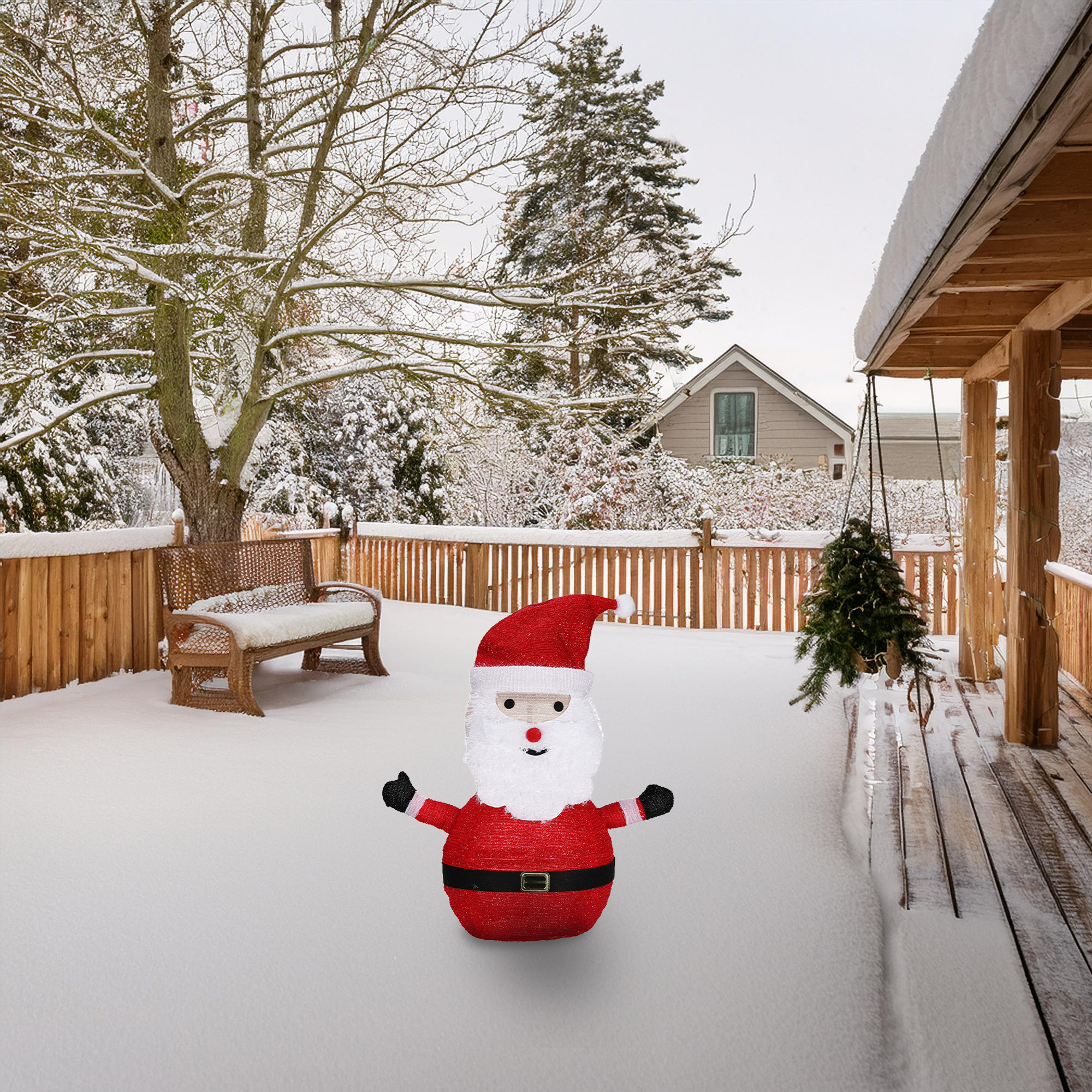 Una escena nevada presenta una decoración de Papá Noel en una cubierta nevada. También se ve un banco, árboles, una casa y un árbol colgante.