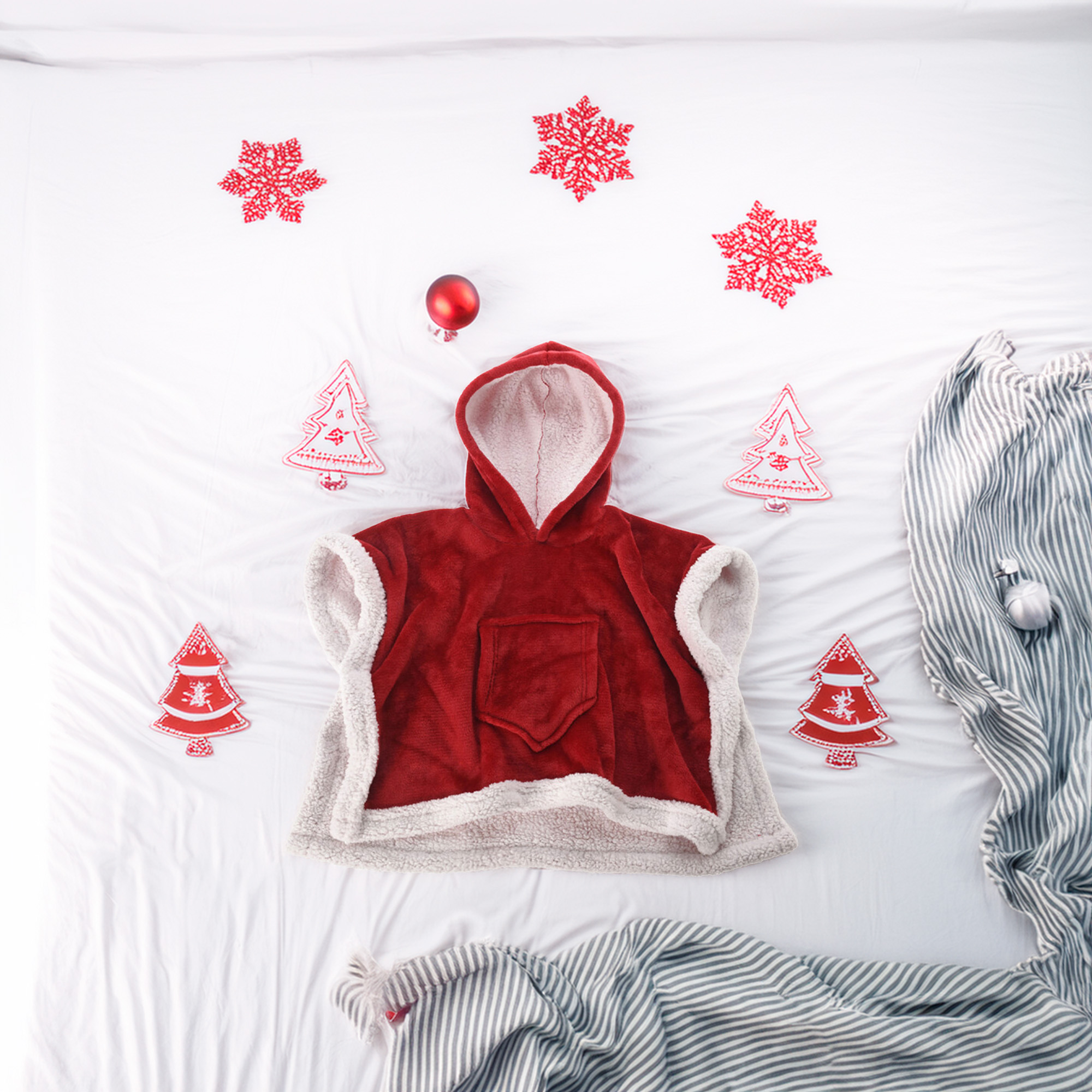 Un poncho rojo con capucha y ribete blanco está sobre una cama blanca, rodeado de adornos de copos de nieve y árboles rojos y mantas a rayas.