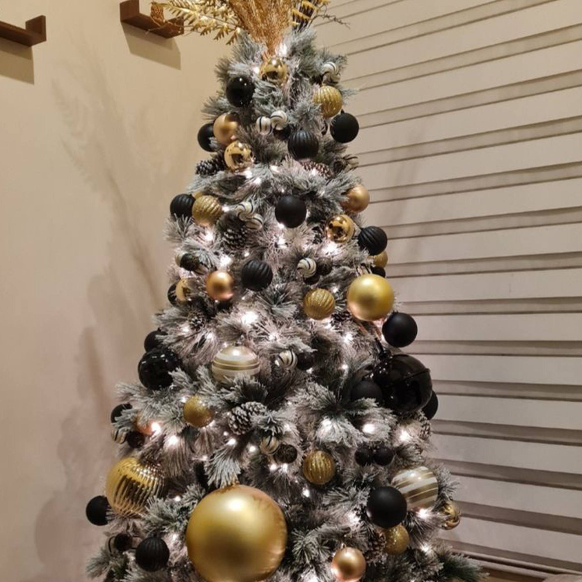 Árbol de Navidad decorado con adornos negros y dorados, y luces blancas. El fondo es una pared y persianas.
