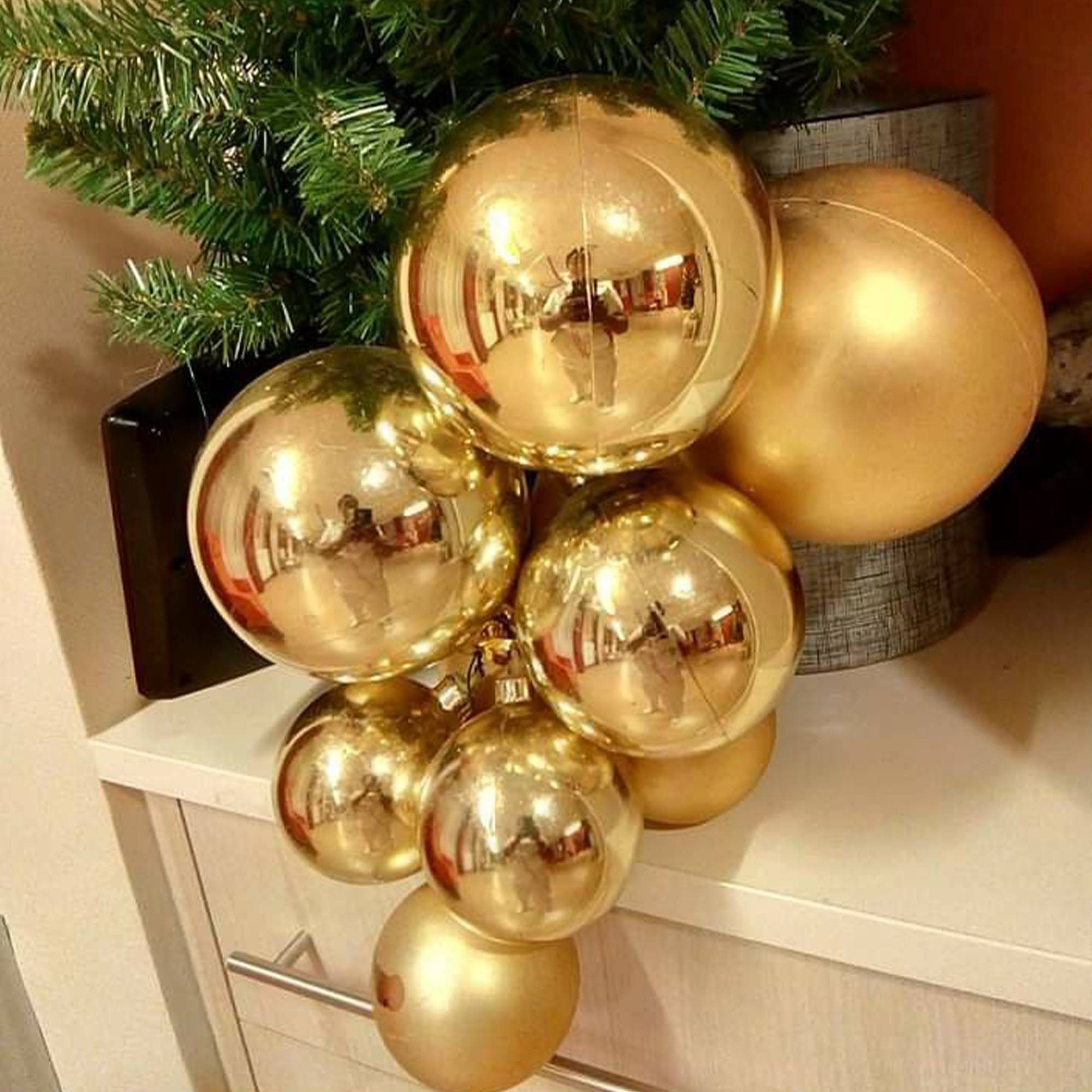 Primer plano de adornos navideños dorados. En el fondo se ven agujas de pino verdes. Algunos adornos reflejan una persona y una habitación.