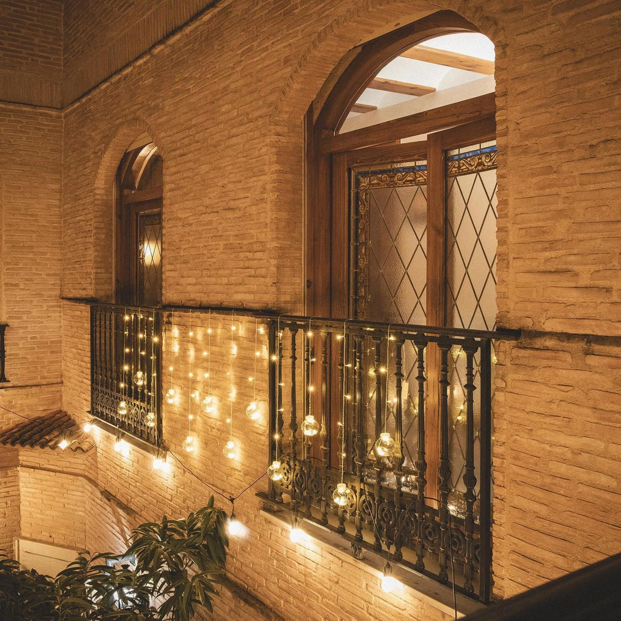 Un edificio de ladrillo con ventanas arqueadas y balcones. Luces de cadena cuelgan del balcón, iluminando la escena.