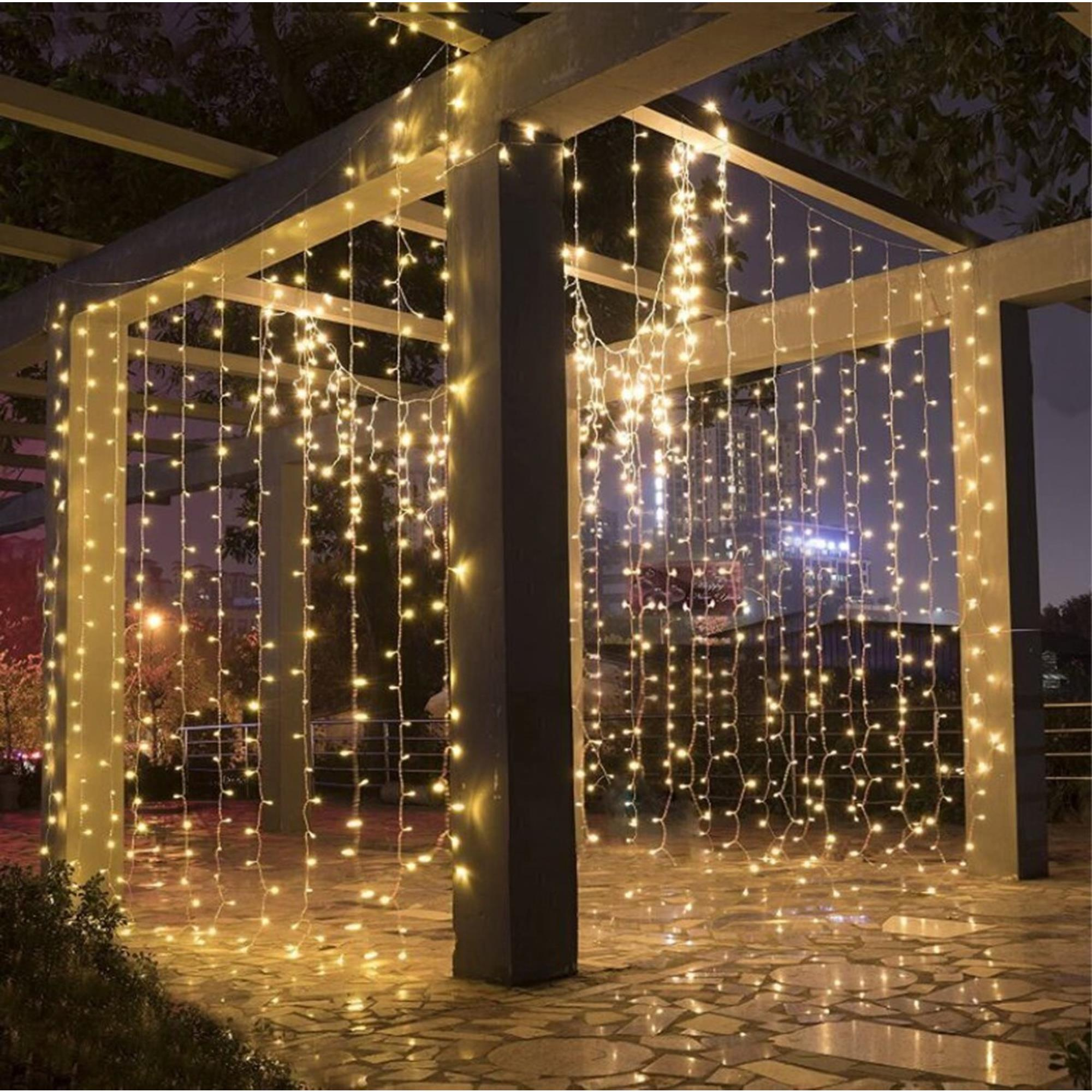 Luces de cadena cuelgan de una pérgola por la noche, iluminando un patio. Luces de la ciudad visibles al fondo.