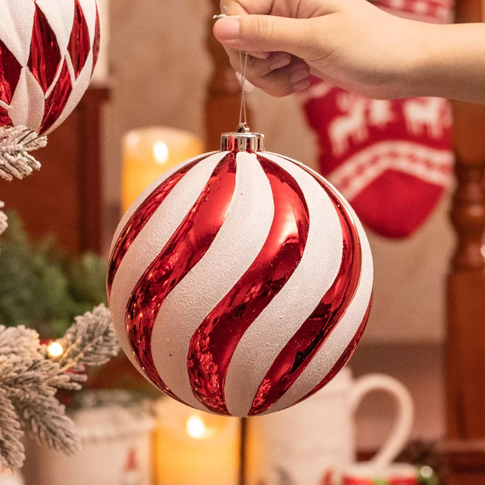 Una mano sostiene un adorno navideño a rayas rojas y blancas. El fondo incluye velas, una media y vegetación.