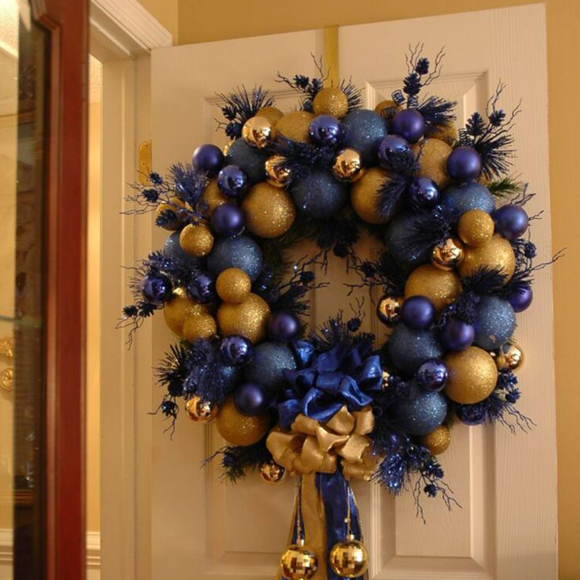 Una corona navideña con adornos azules y dorados cuelga de una puerta blanca. Un lazo dorado y una cinta azul en la parte inferior.