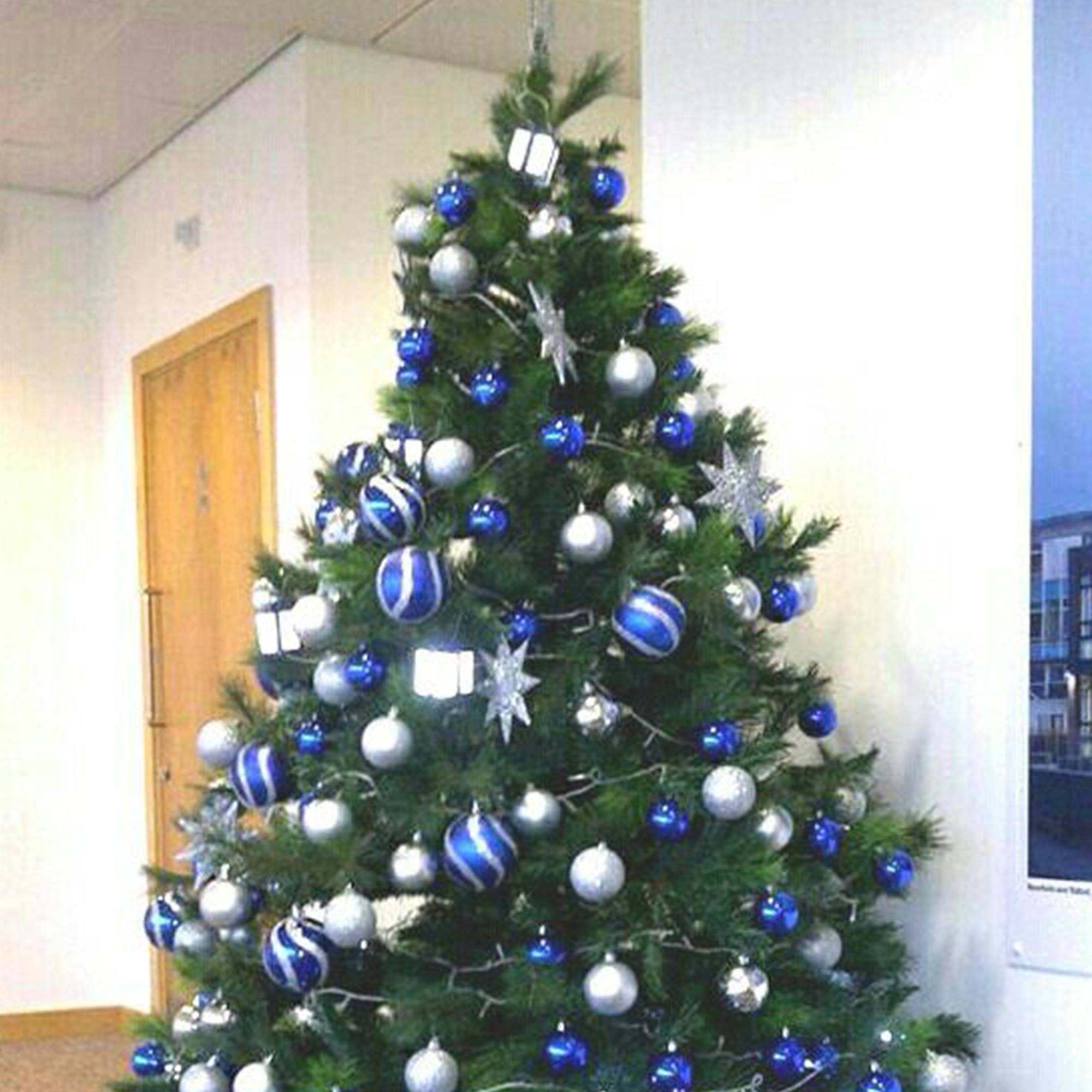 Un árbol de Navidad decorado con adornos azules y plateados. Está en un entorno de oficina cerca de una puerta.