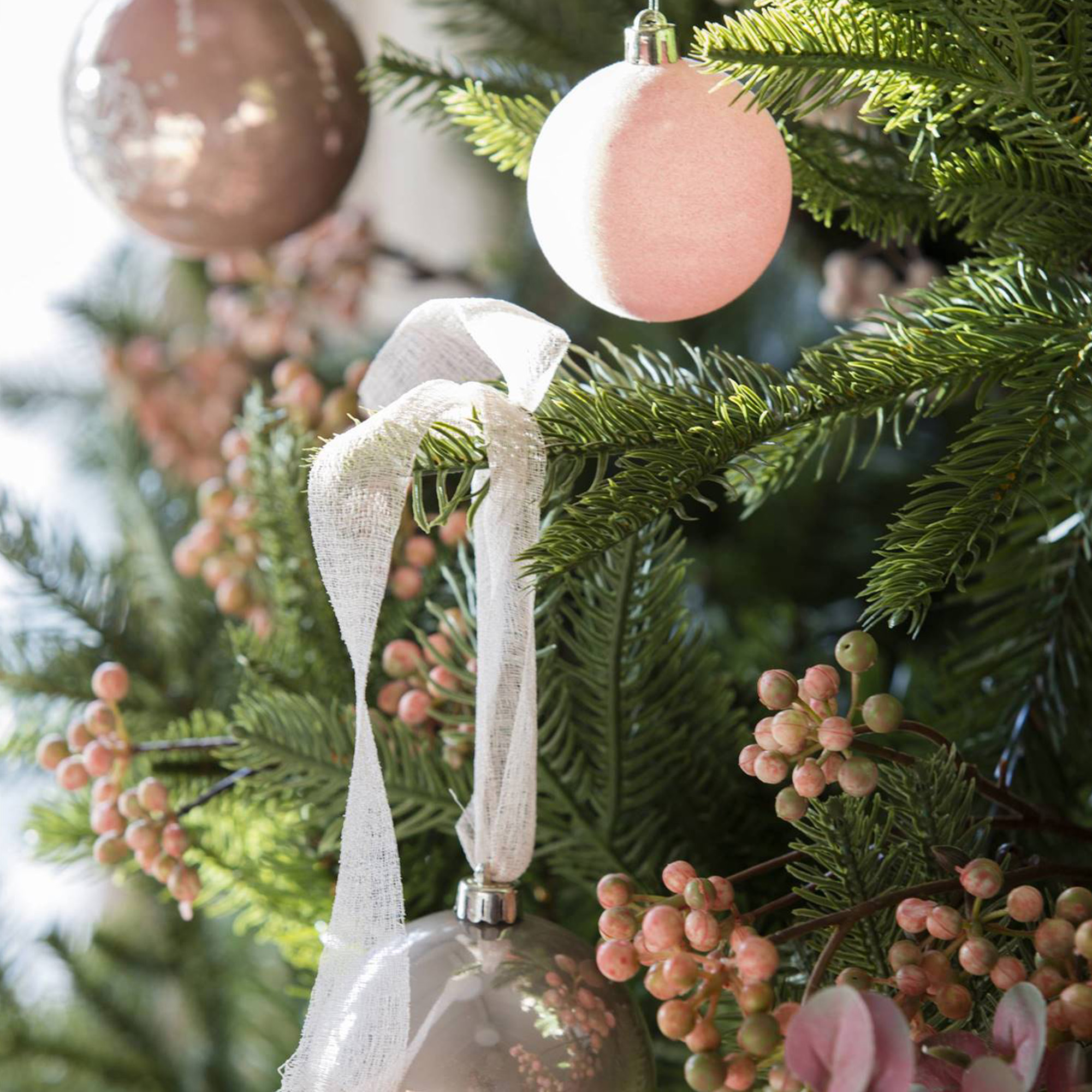 Un árbol de Navidad con adornos y bayas. Adornos rosas y plateados cuelgan de las ramas verdes. También hay cinta blanca.