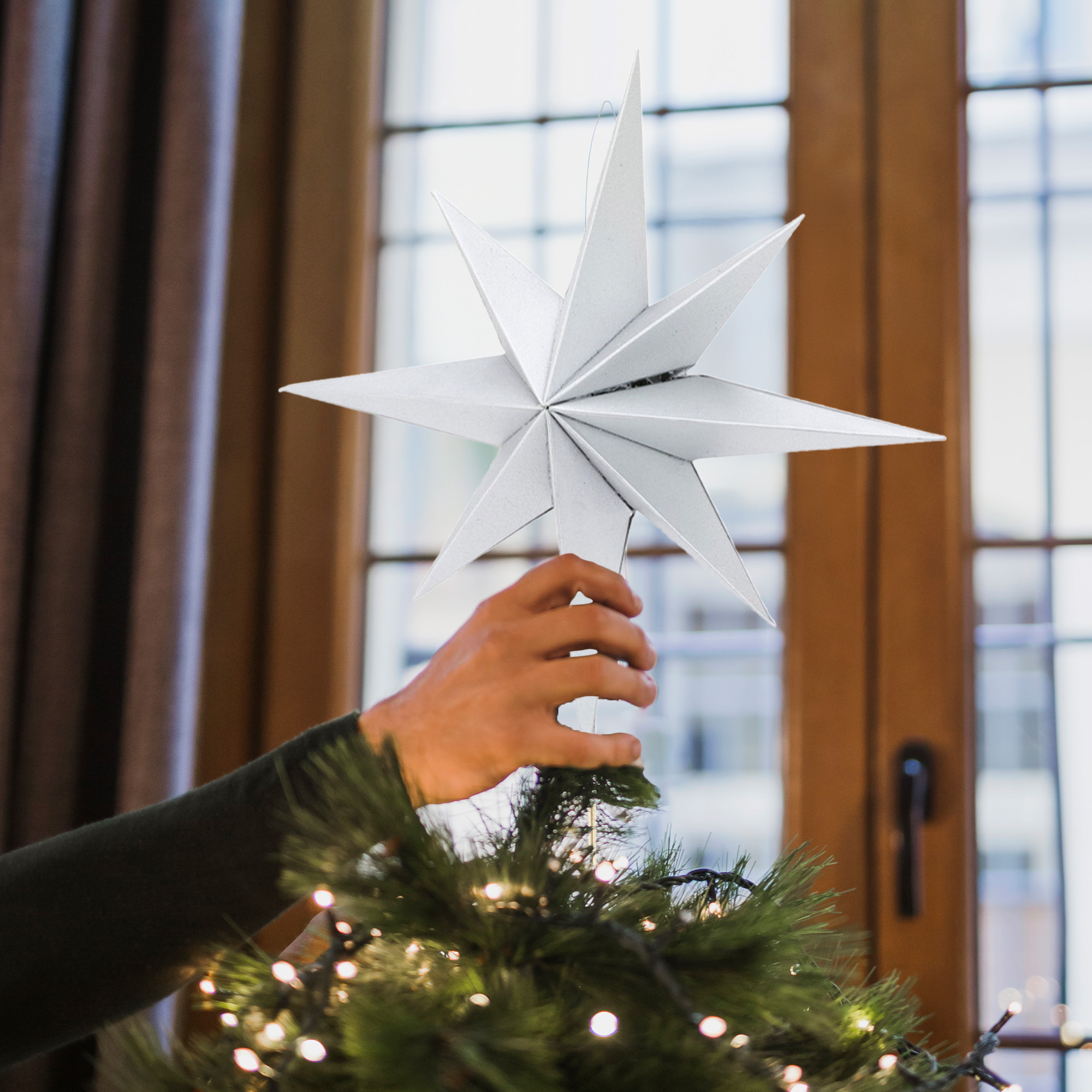 Una mano coloca una estrella blanca en la parte superior de un árbol de Navidad. Luces en el árbol. Ventana al fondo.