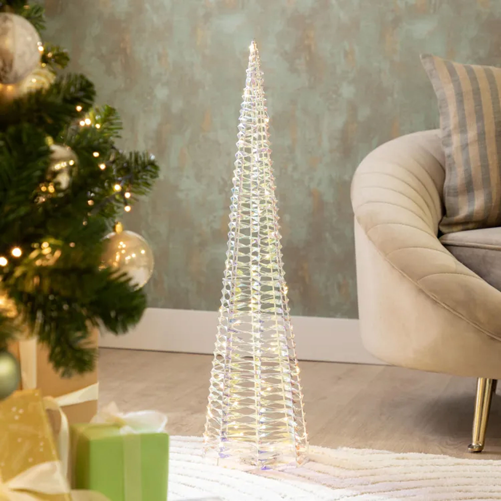 Un árbol de Navidad de cristal iluminado decorativo se encuentra cerca de un árbol decorado, regalos y un sillón beige.