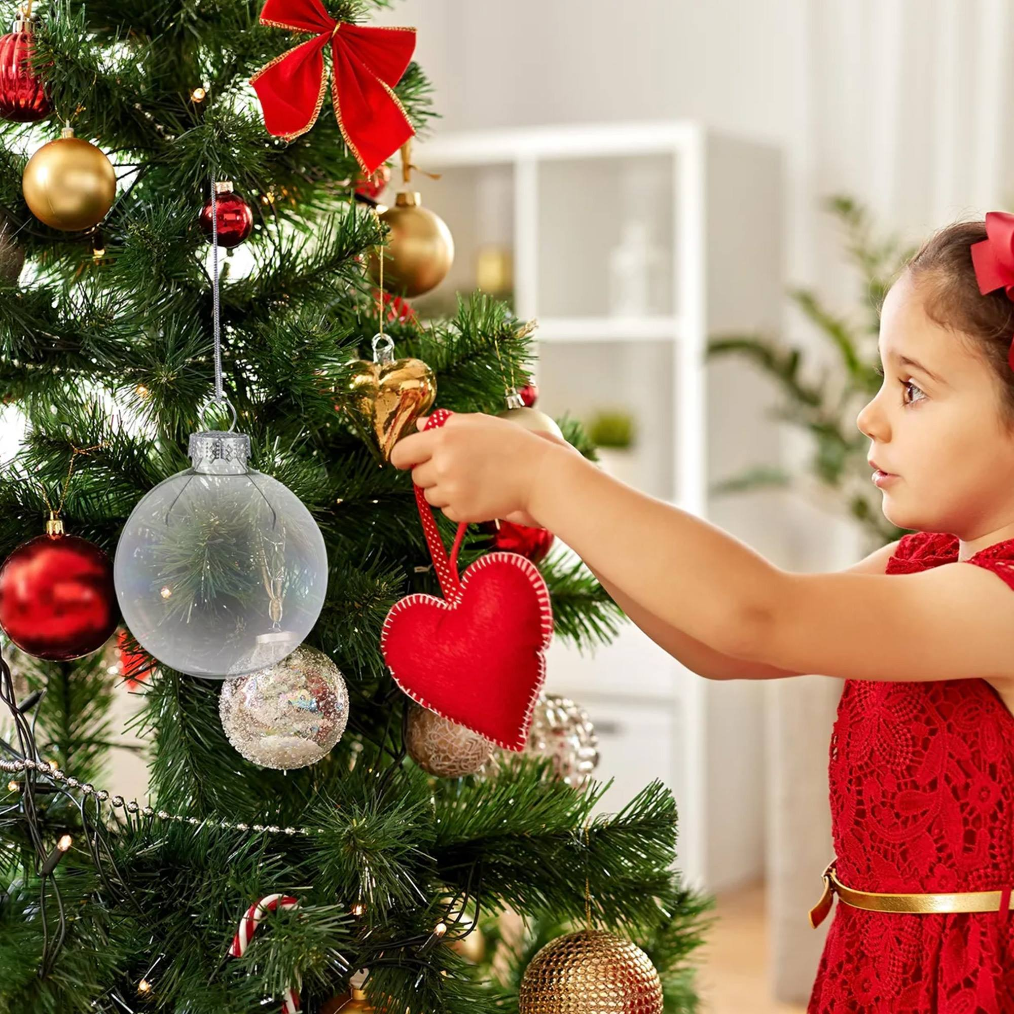 Una niña con un vestido rojo decora un árbol de Navidad con adornos de corazón rojo. El árbol tiene decoraciones doradas y rojas.