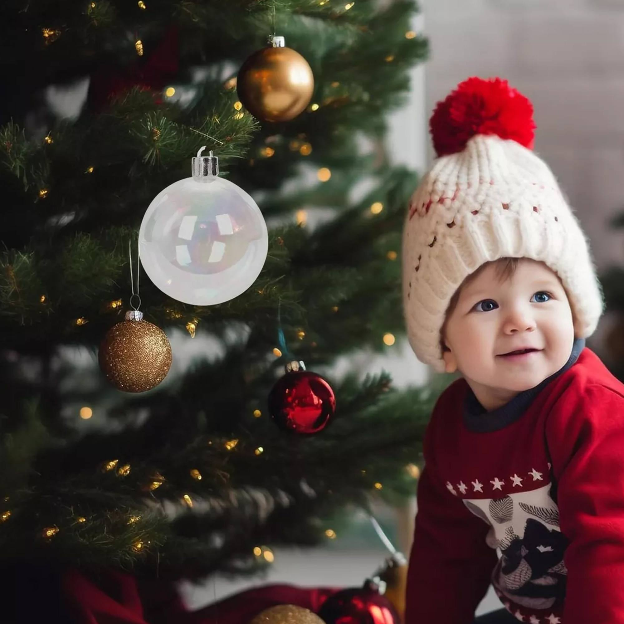 Un bebé con gorro de punto y suéter sonríe cerca de un árbol de Navidad decorado con adornos y luces.