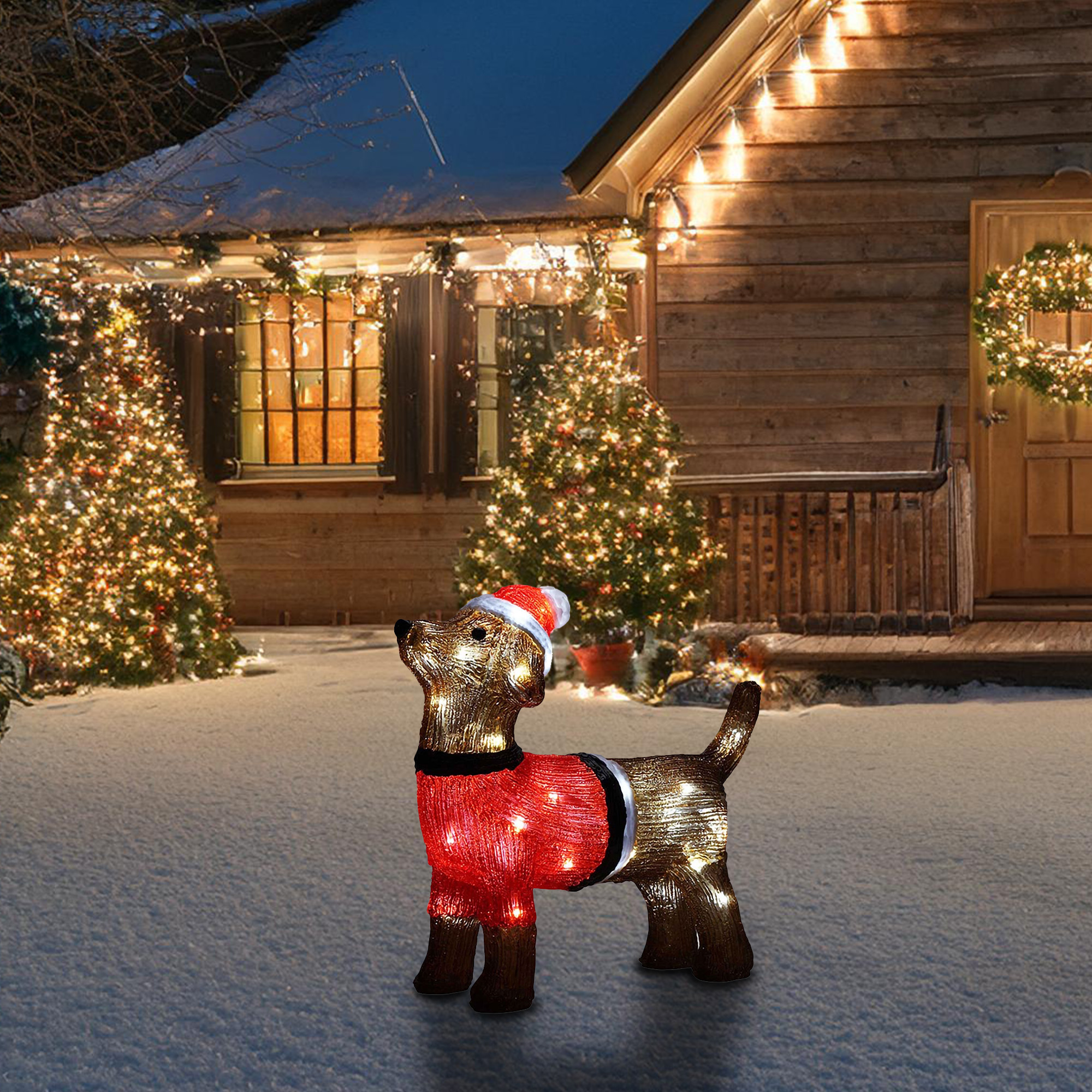 Estatua de perro iluminada con gorro y suéter de Papá Noel en la nieve. Luces navideñas decoran casa y árboles al fondo.