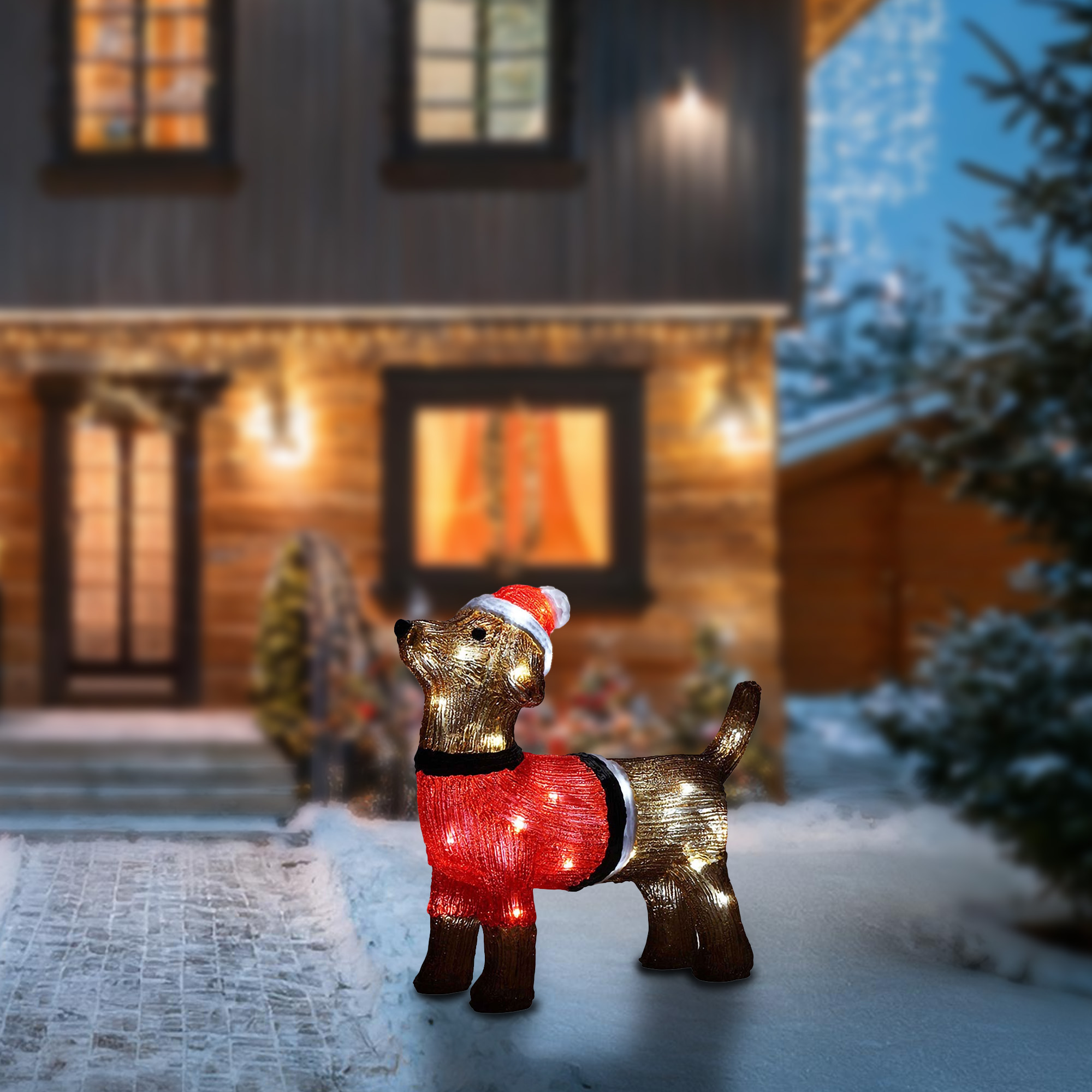 Una decoración navideña de un perro con gorro de Papá Noel y suéter rojo está en la nieve. Una casa al fondo.