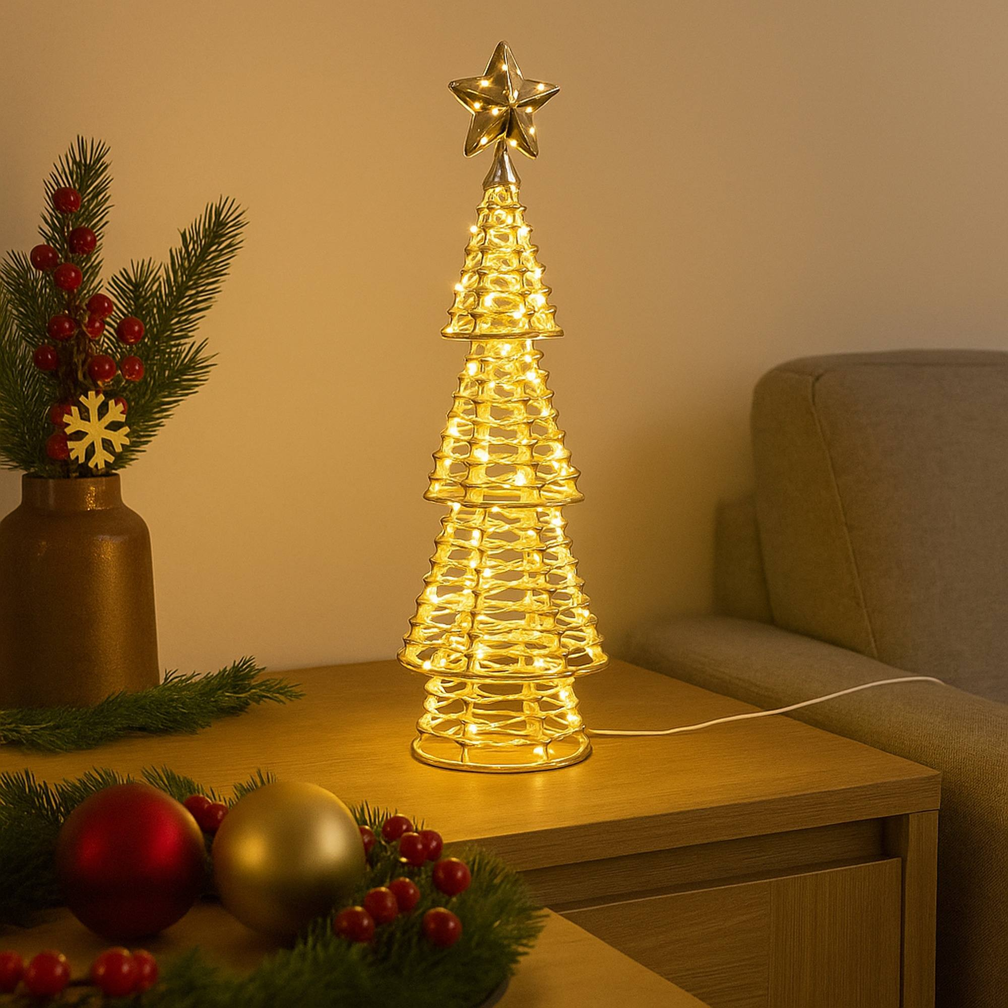Lámpara en forma de árbol de Navidad sobre una mesa con adornos, un jarrón con bayas y un sofá. Iluminación cálida.