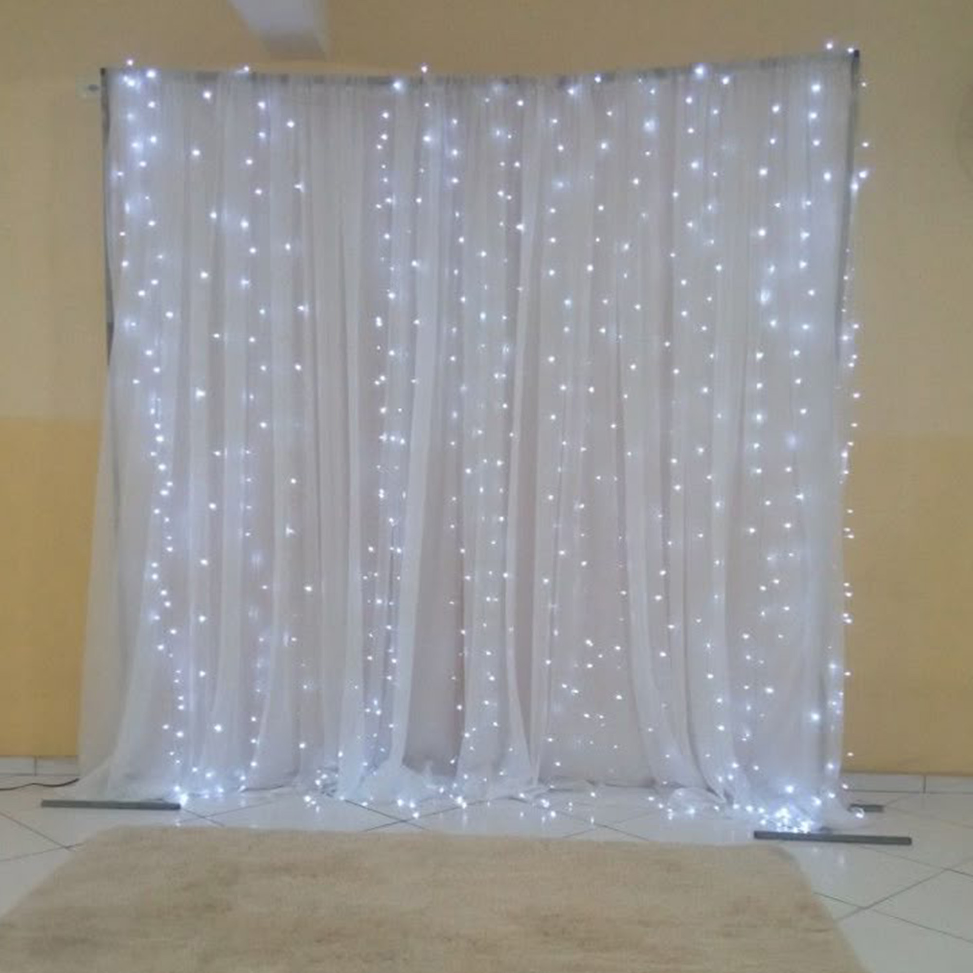 Cortina blanca con luces de cadena forma un telón de fondo. Una alfombra beige está al frente. La pared es amarillo claro.