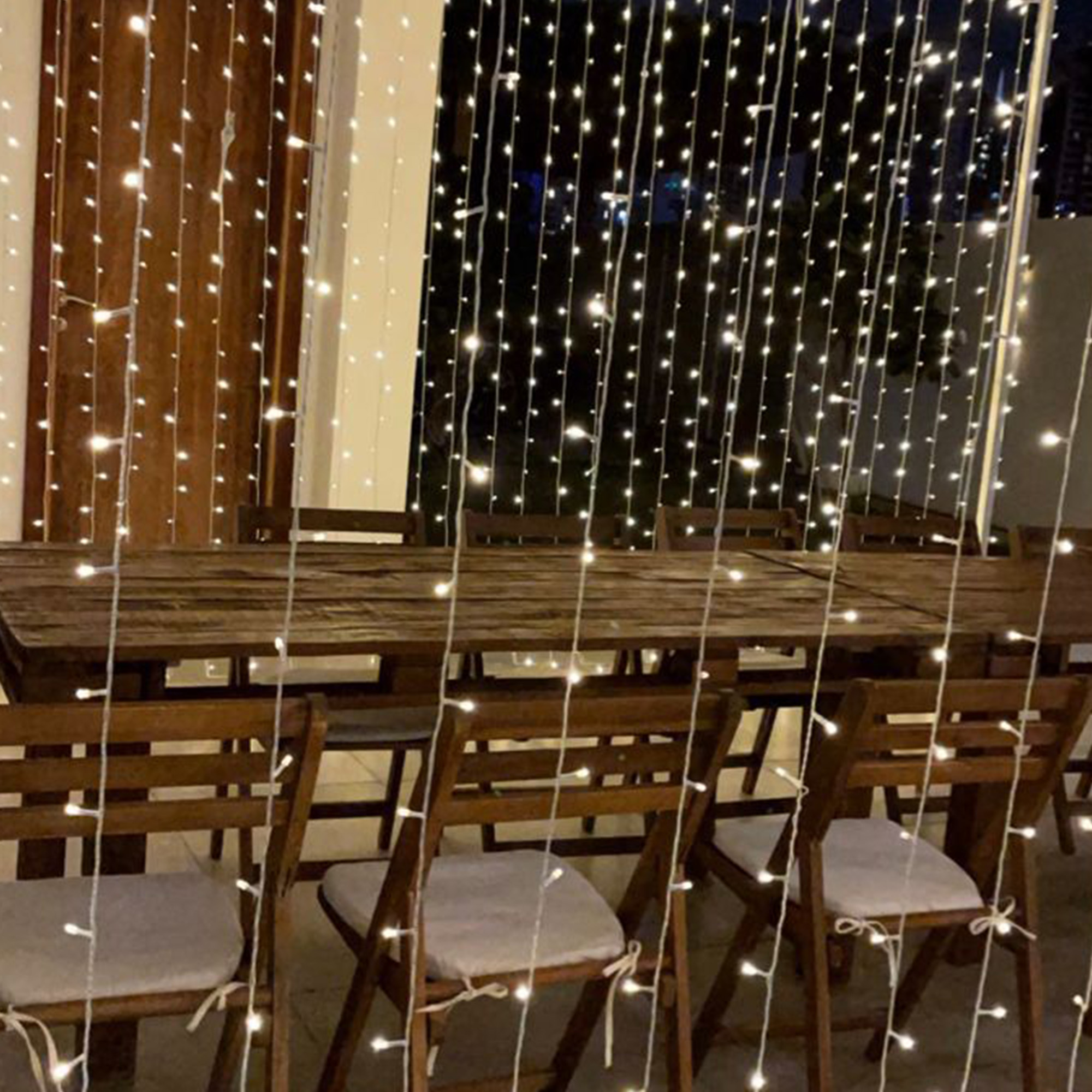 Una mesa y sillas de madera están preparadas, con luces de cadena en el fondo, creando un ambiente cálido y acogedor.