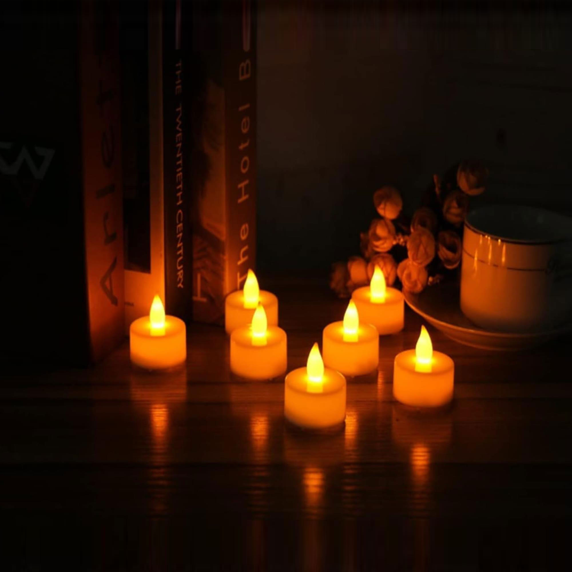 Varias velas amarillas pequeñas y encendidas sobre una superficie de madera. Libros y una taza al fondo. Escena tenue.