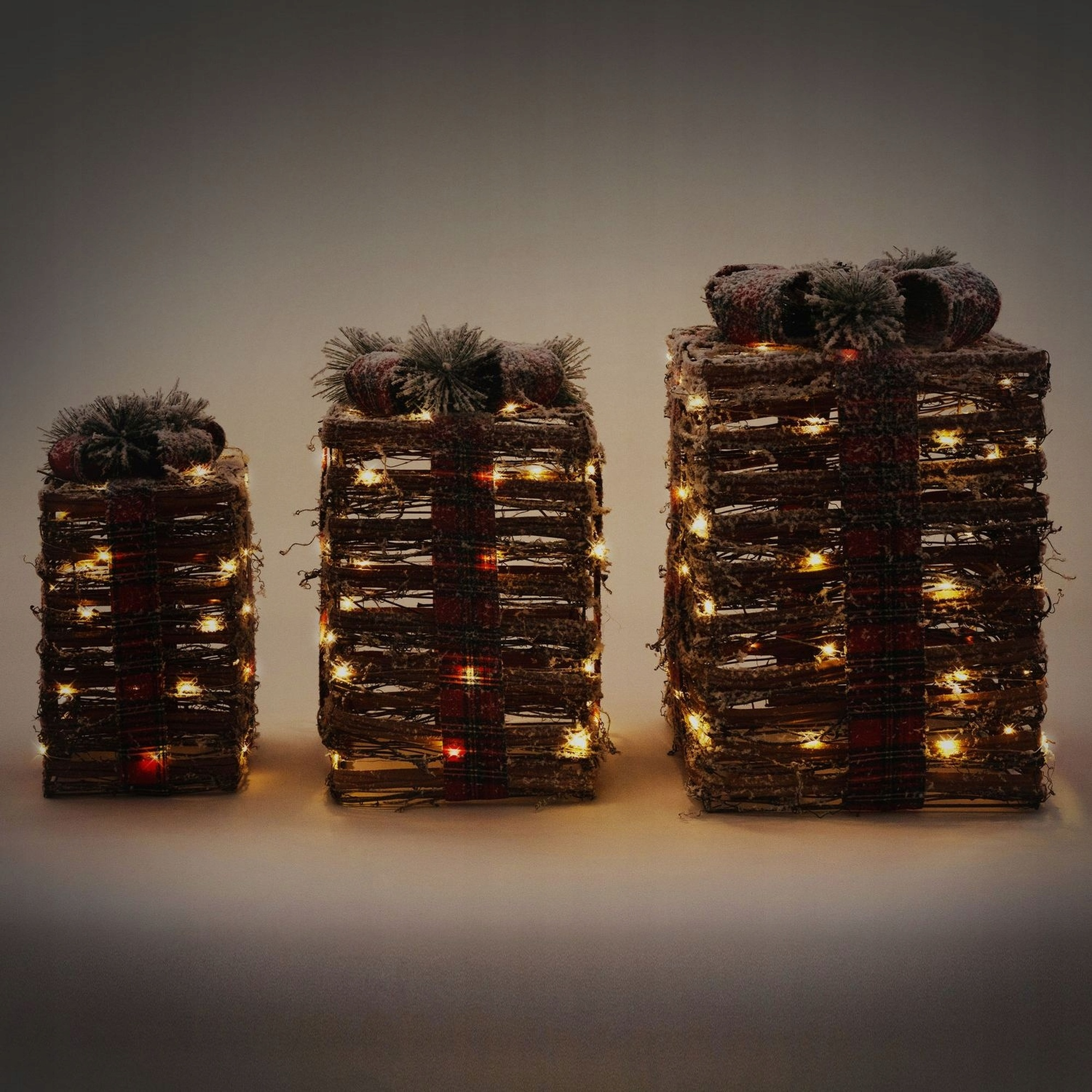 Tres decoraciones de cajas de regalo iluminadas. Envueltas en material tejido marrón y cinta de cuadros rojos, con lazos cubiertos de nieve.